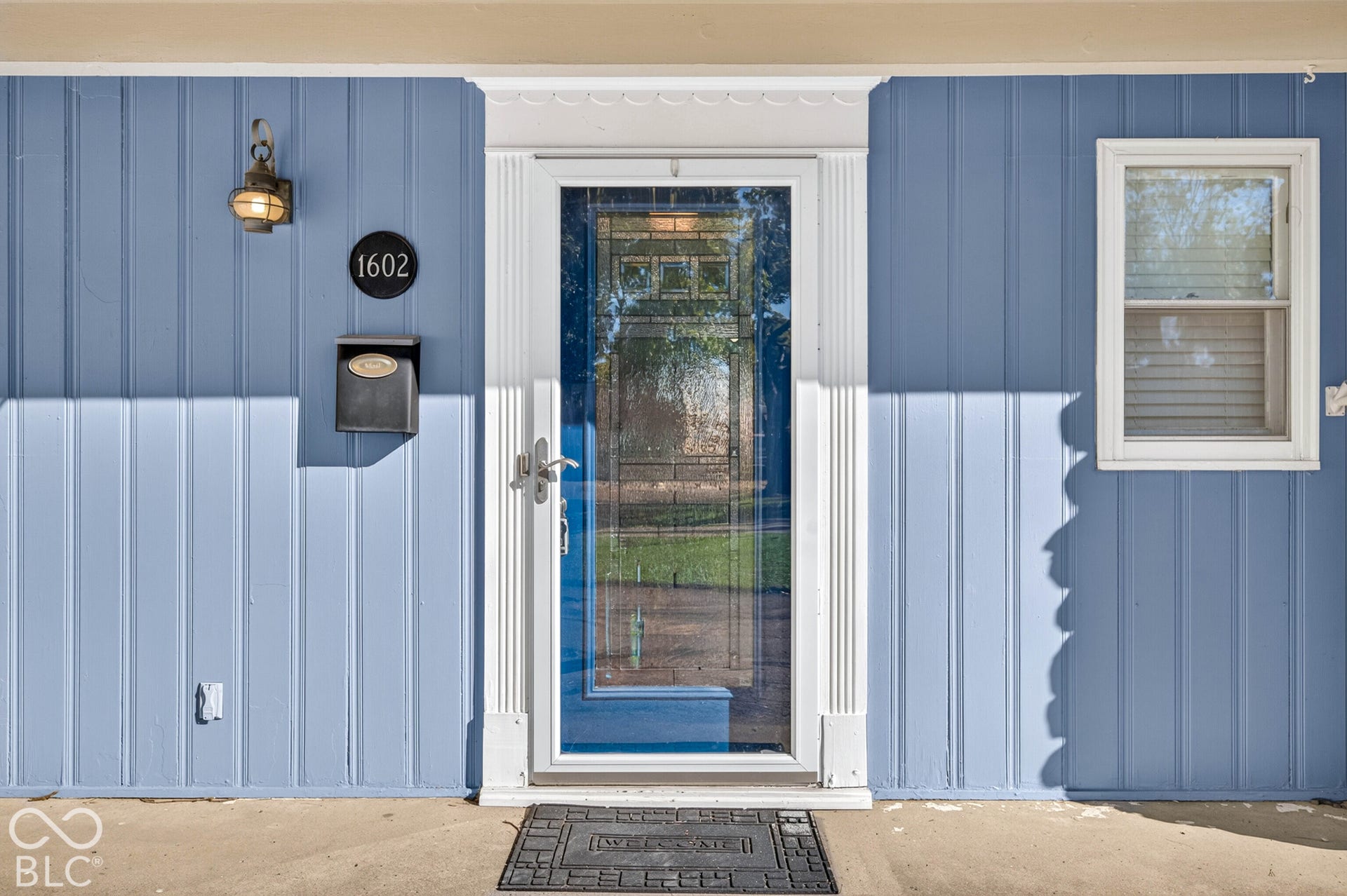 Board and batten exterior with a storm door, an outdoor wall light, and a window with blinds