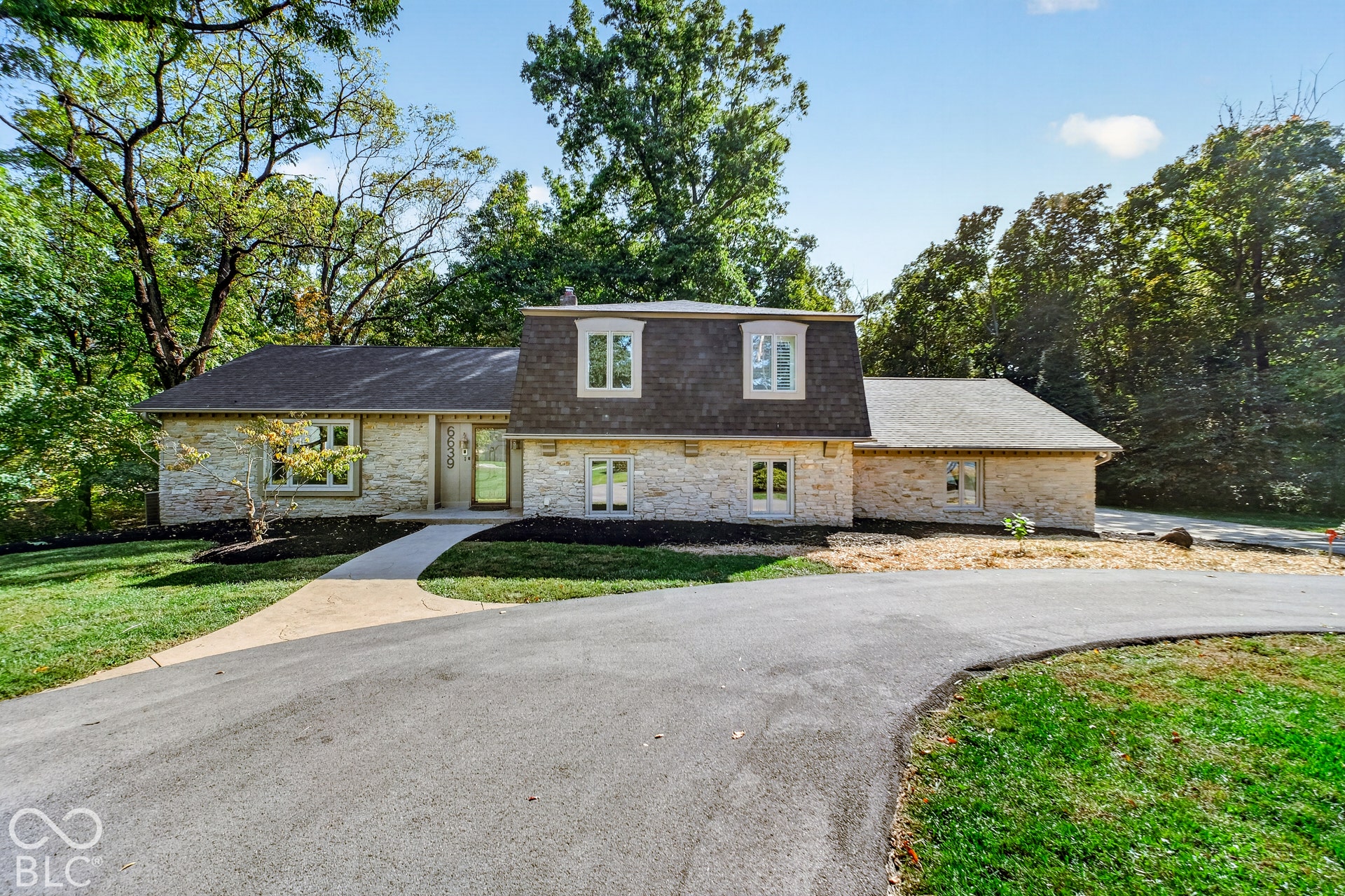 Stone exterior with dormer windows, asphalt driveway, and mature landscaping