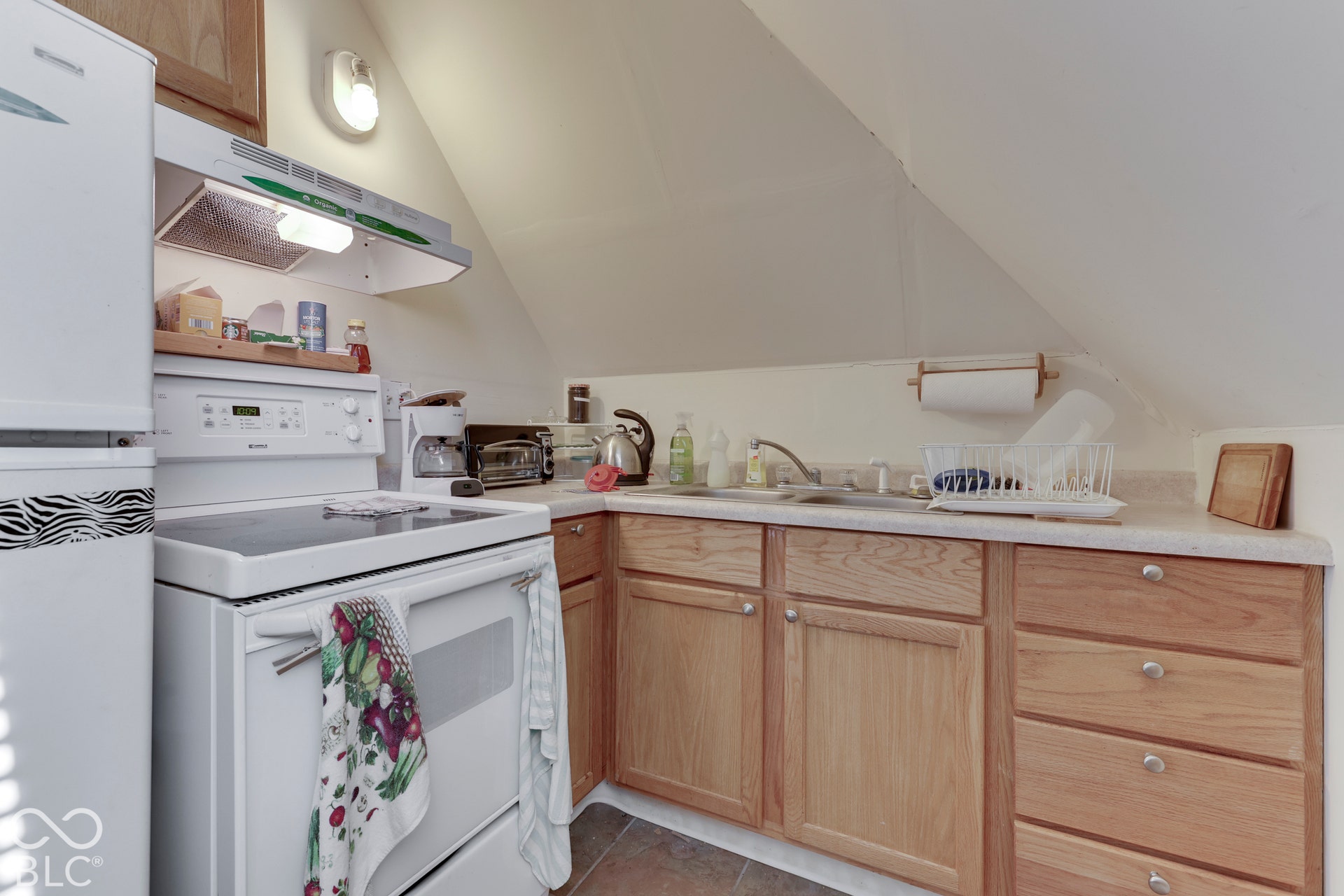White electric range with an overhead exhaust fan, wood-finish cabinetry, light-toned countertops, and a single-basin sink