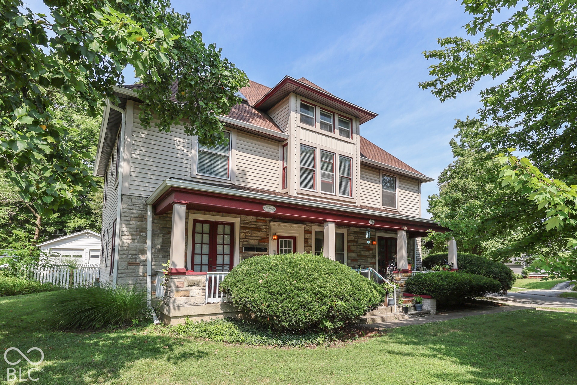 Historic two-story residence featuring stone and siding exterior, expansive covered porch, multiple windows, and mature landscaping.