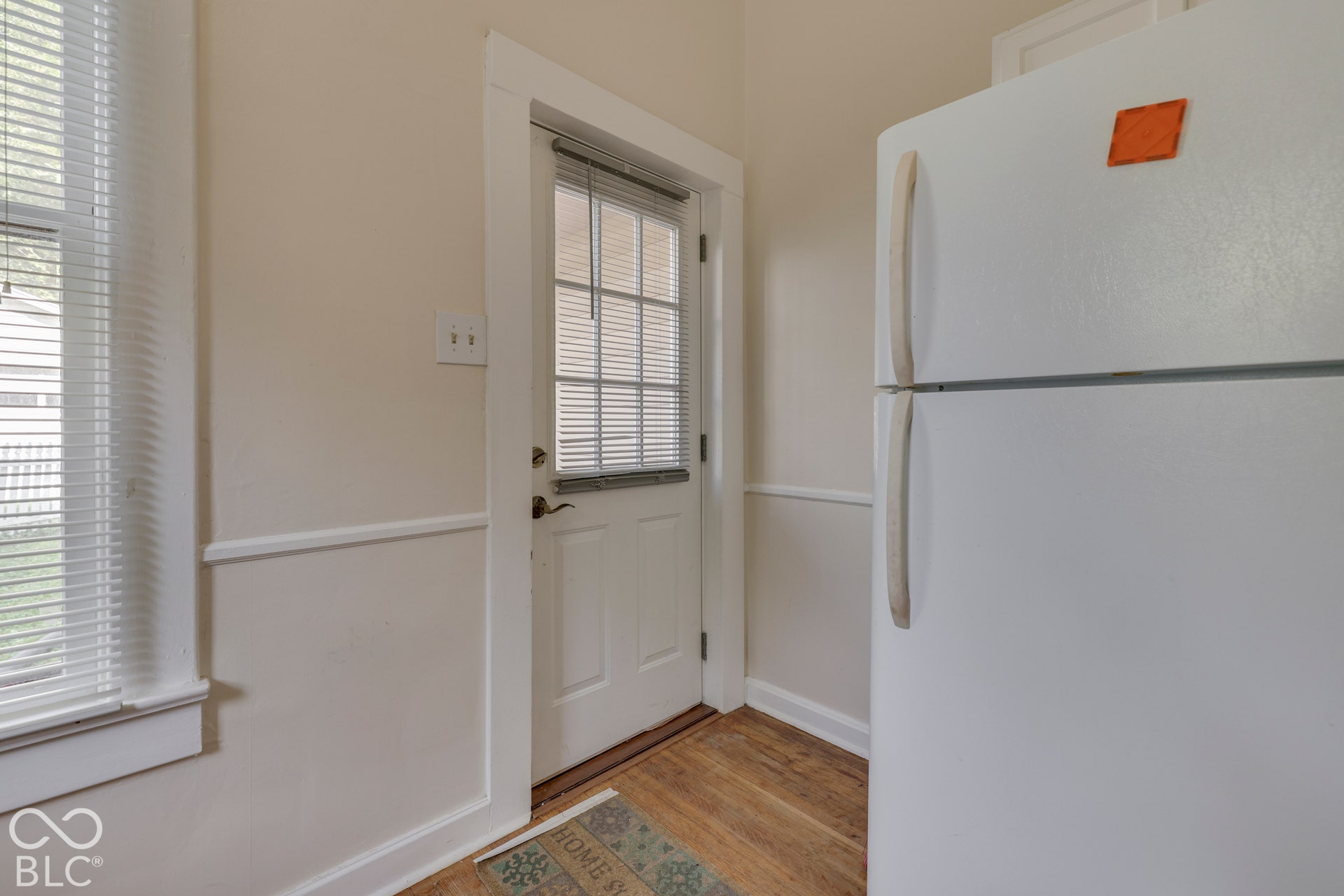 Unit 1 White full-size refrigerator, wood-finish flooring, white paneled door with glass insert, white window with horizontal blinds, and neutral wall paint