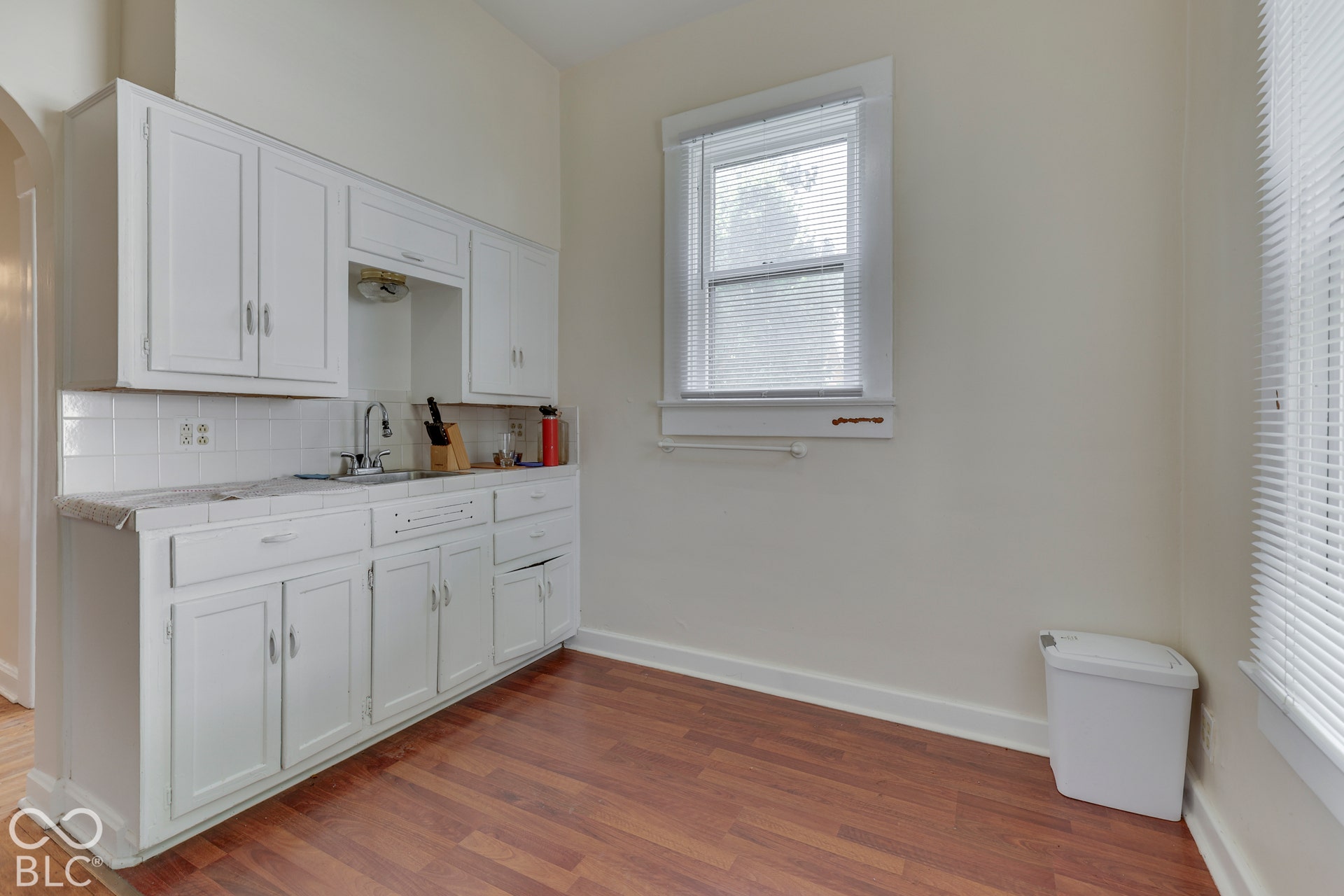 Unit 1 White cabinetry with chrome hardware, white tile backsplash, stainless steel sink, wood-finish flooring, and natural light from a window