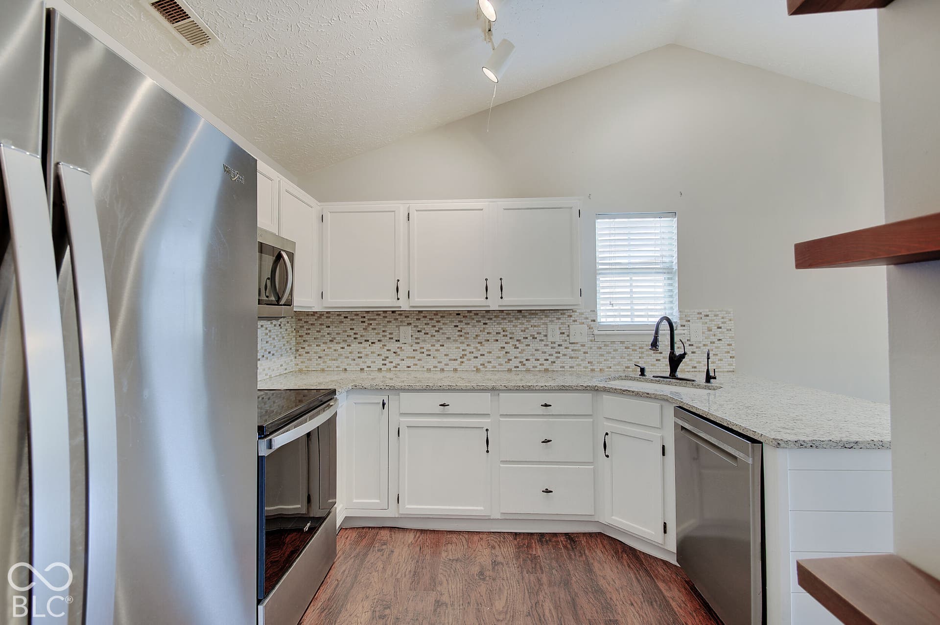 Vaulted ceiling kitchen with white cabinetry, stainless steel appliances, speckled stone countertops, and wood-finish flooring