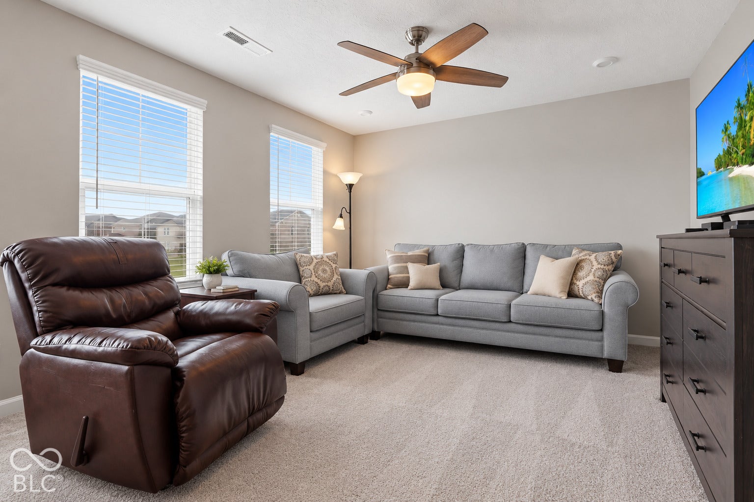 Soft gray wall color, light-toned carpet, two windows with blinds, ceiling fan with integrated lighting, and recessed ceiling light