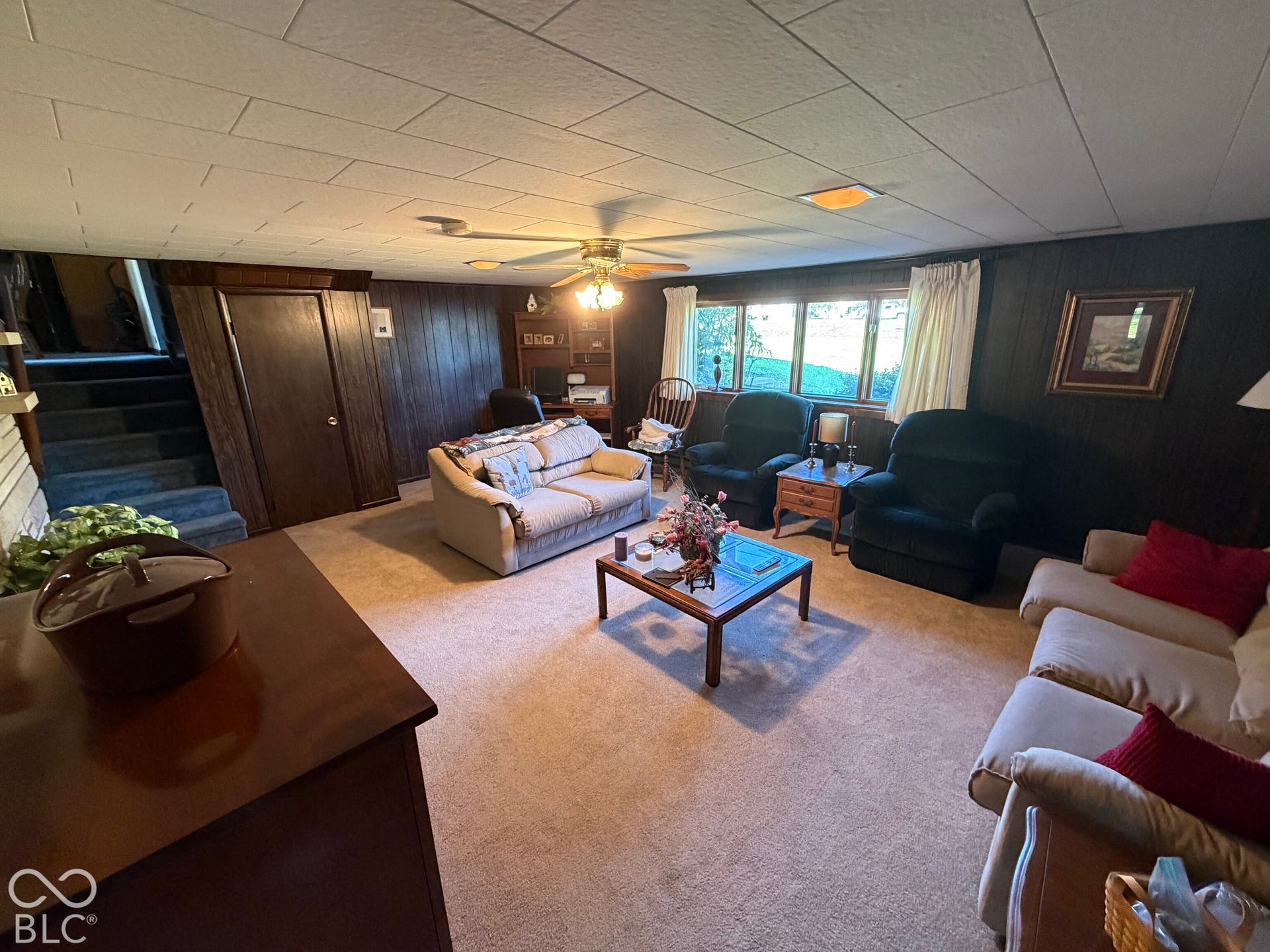 Window-lined wall, ceiling fan with integrated lighting, wood paneling, carpeted flooring, and built-in staircase
