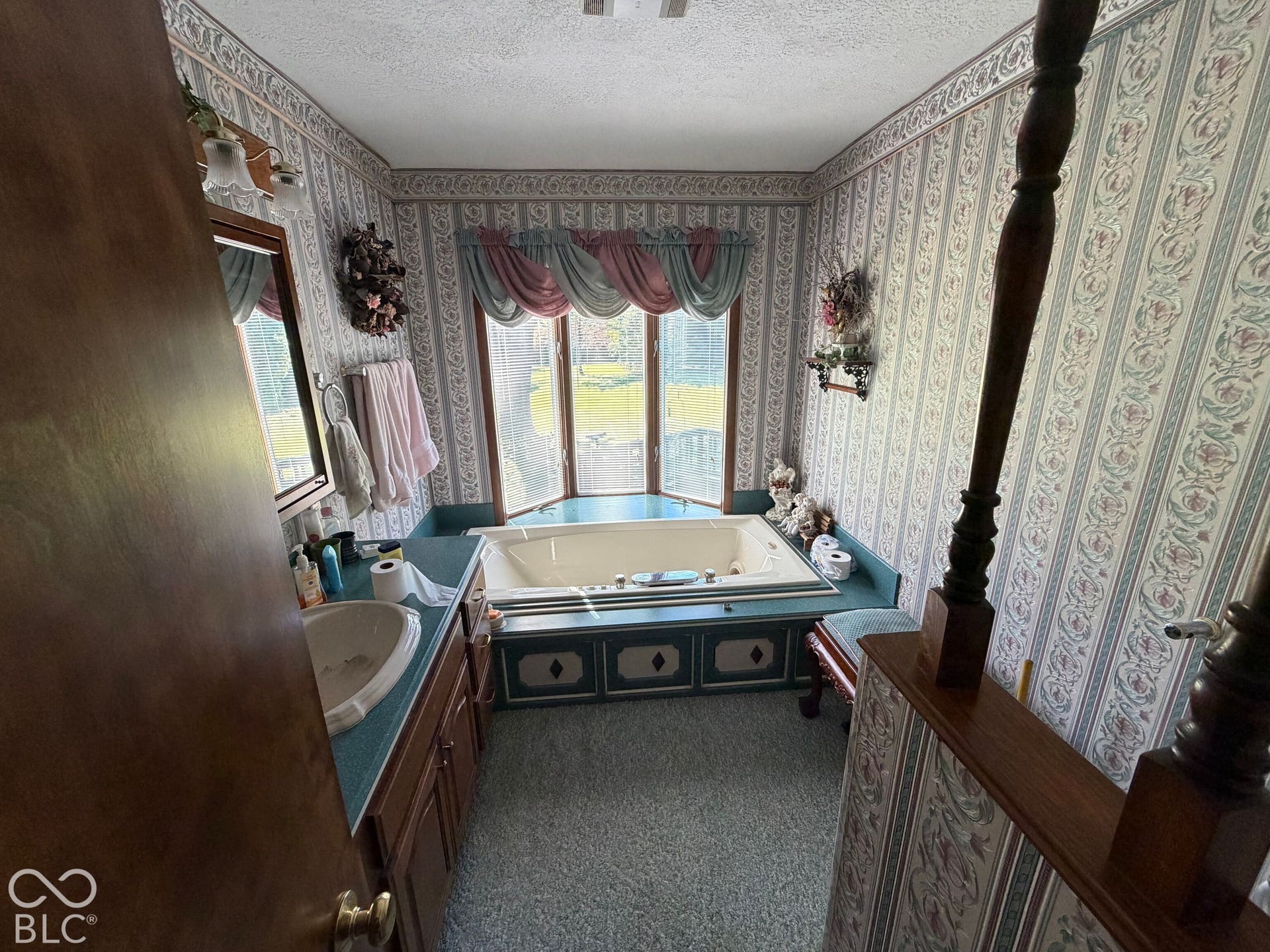 Built-in soaking tub with a bay window, wood-finish vanity with an integrated sink, and patterned wallpaper