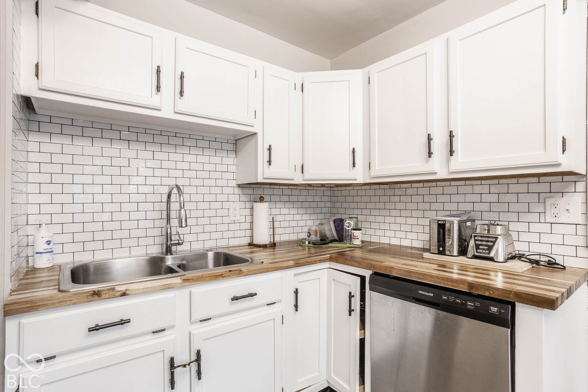 White cabinetry with dark hardware, subway tile backsplash, wood-finish countertops, stainless steel double basin sink, and an integrated stainless steel dishwasher