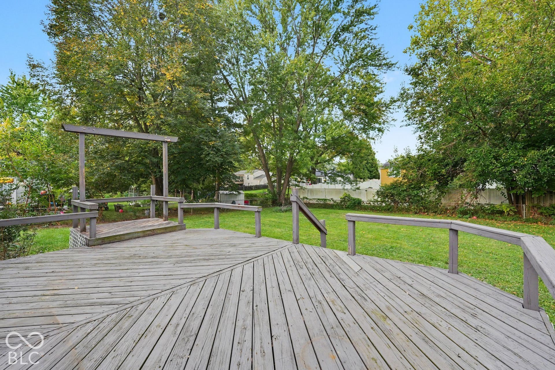 Wood deck with railing, featuring a raised platform, overlooking a grass lawn with mature trees