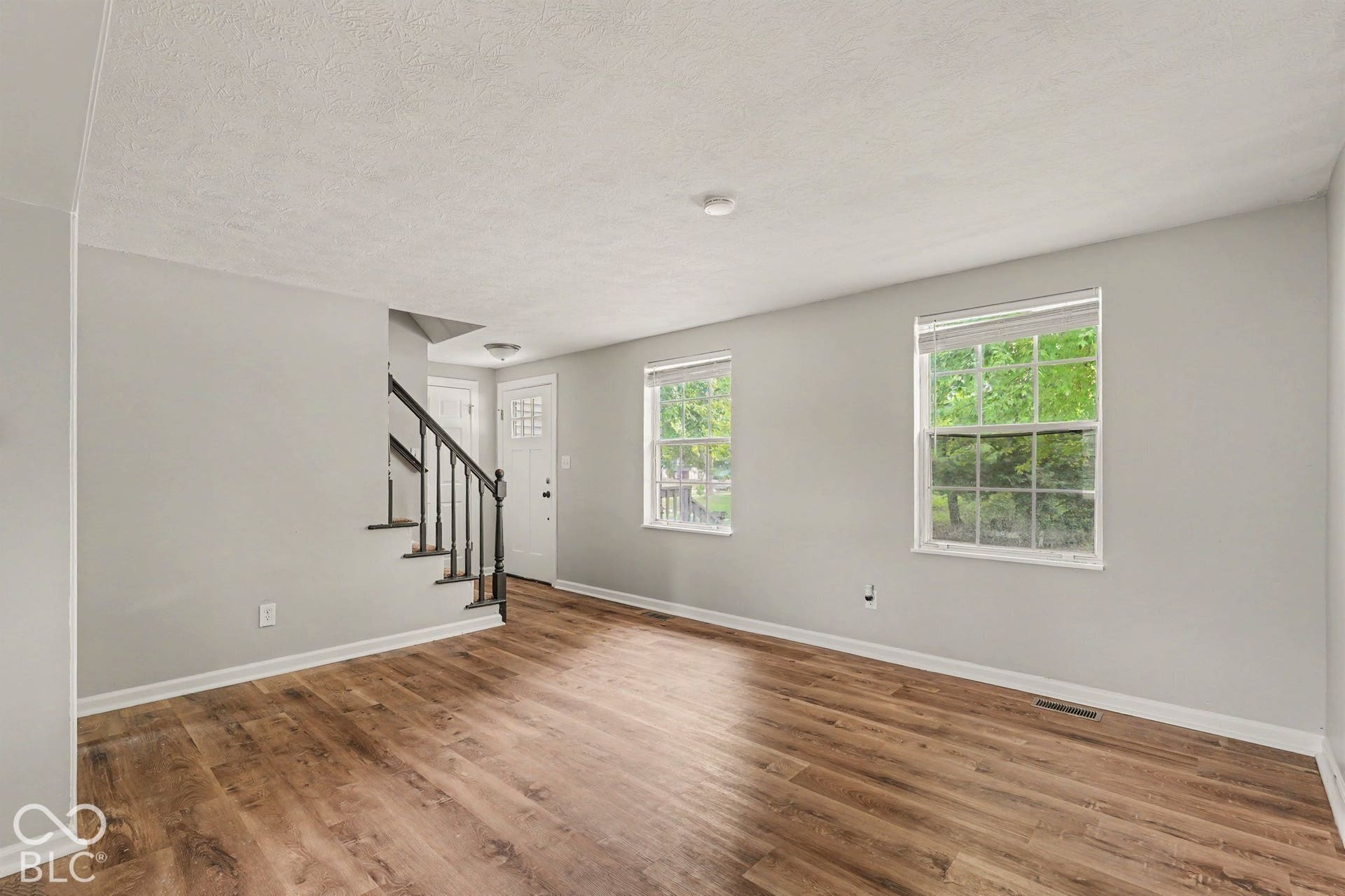 Wood-finish flooring, two large windows, white walls, black stair railing, and a white front door with decorative lites