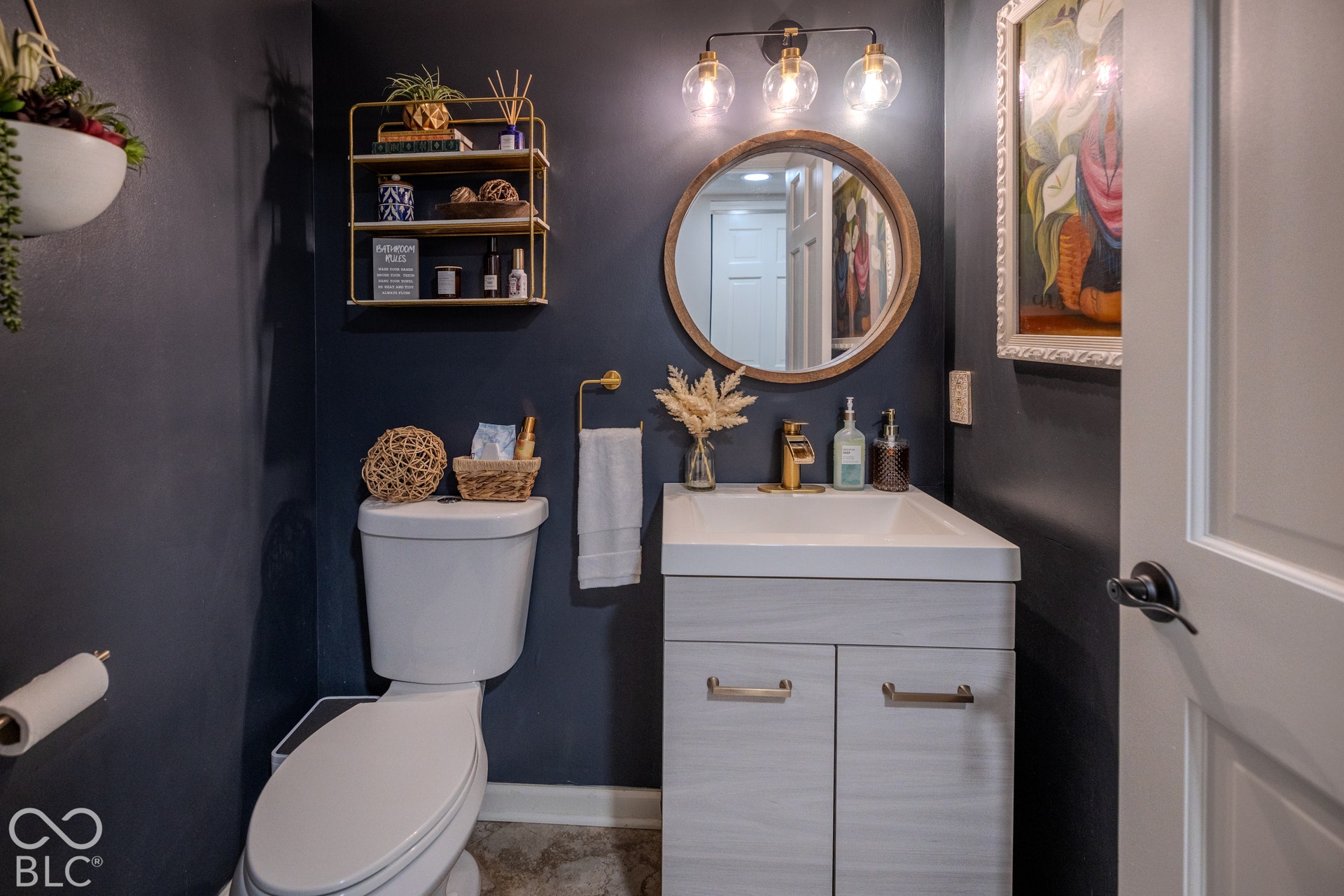 6416 Manchester Drive, Fishers, IN 46038 Stylish powder room featuring a white vanity with dual drawers and brushed brass hardware, white vessel sink, and dark navy walls with natural wood-framed round mirror.