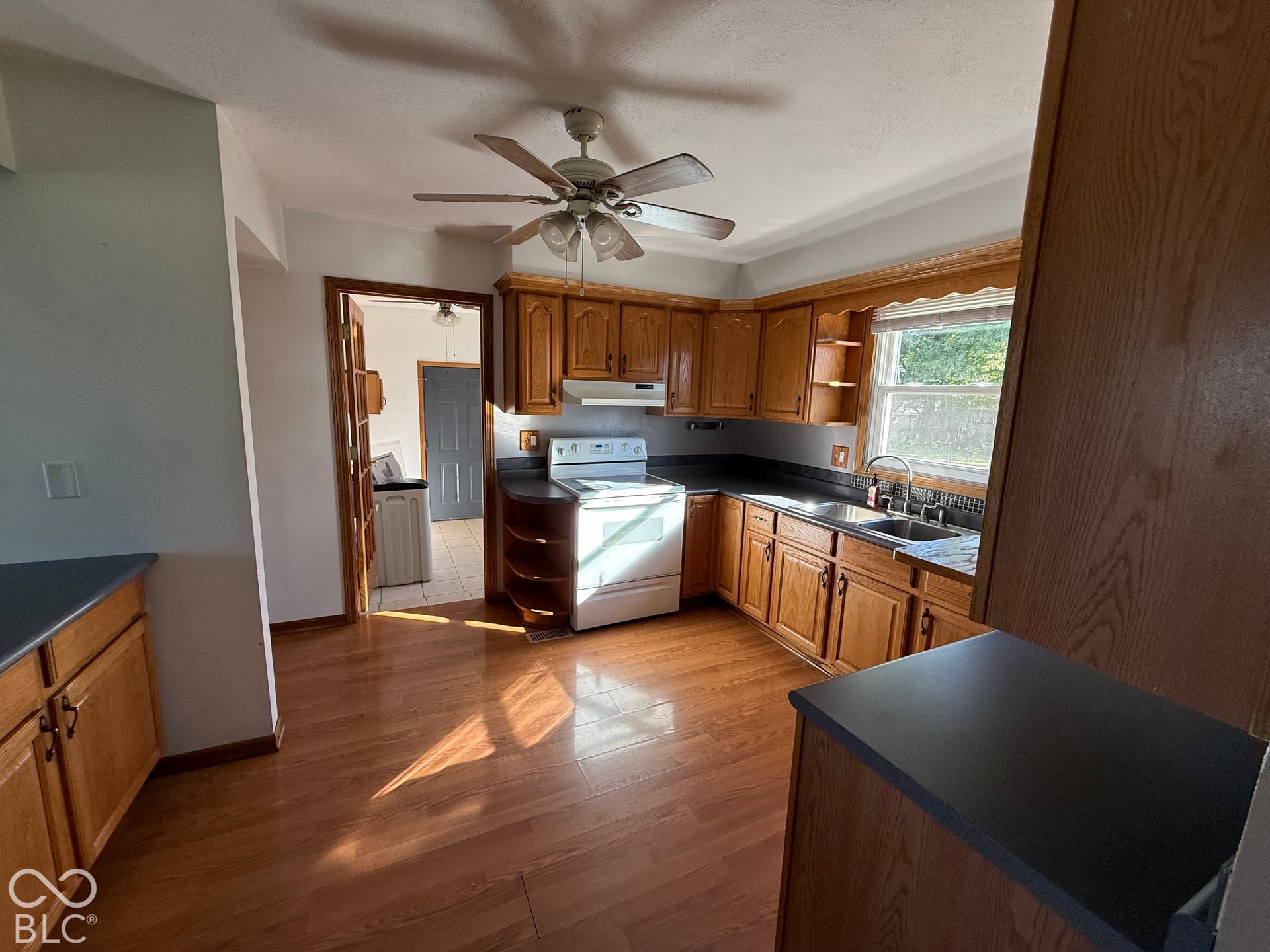 Wood-finish flooring throughout, wood cabinetry, dark countertops, ceiling fan with light fixture, and a stainless steel sink with a high-arc faucet