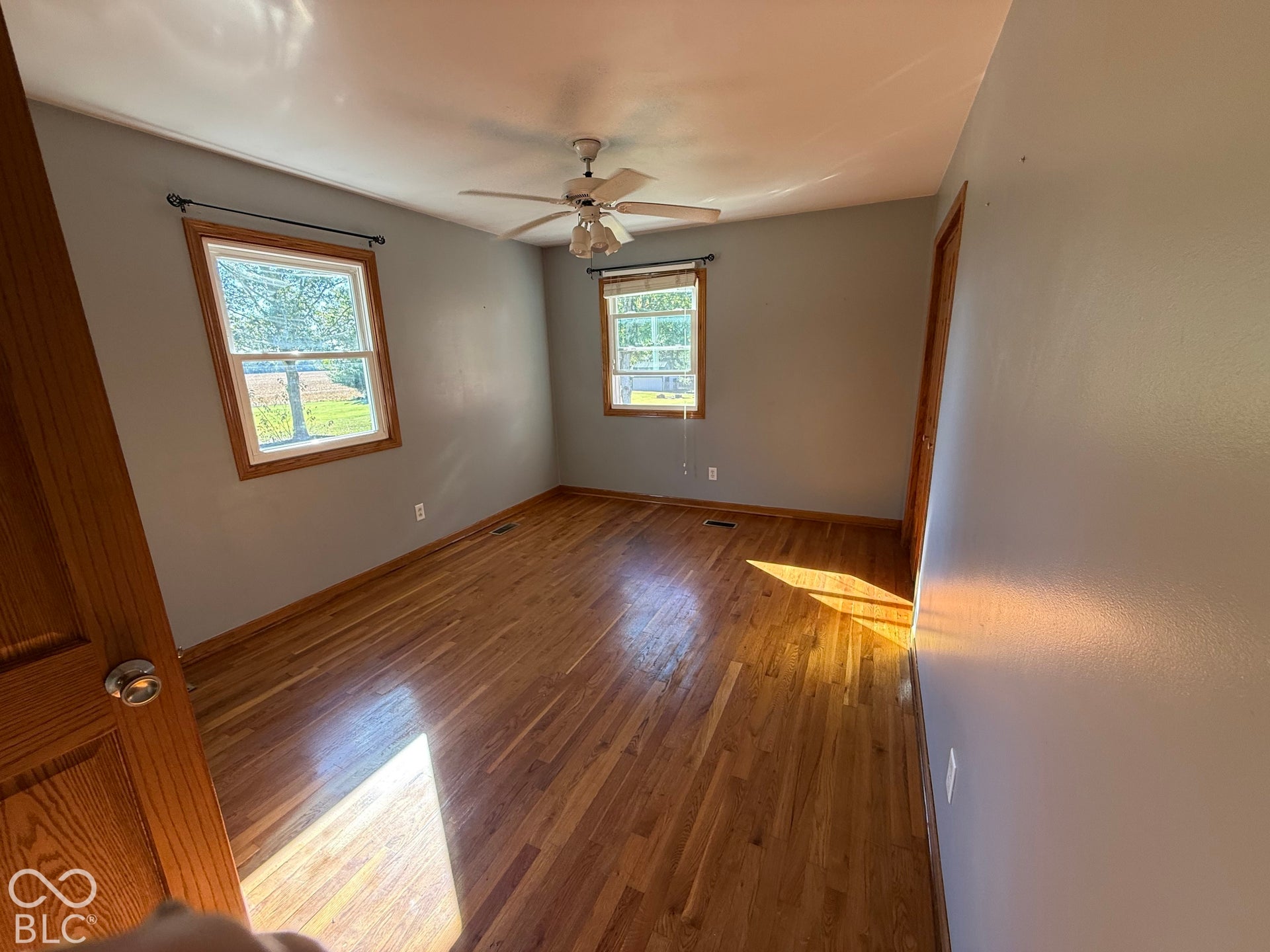 Wood-finish flooring, two windows with wood trim, ceiling fan light fixture, and wood-panel door