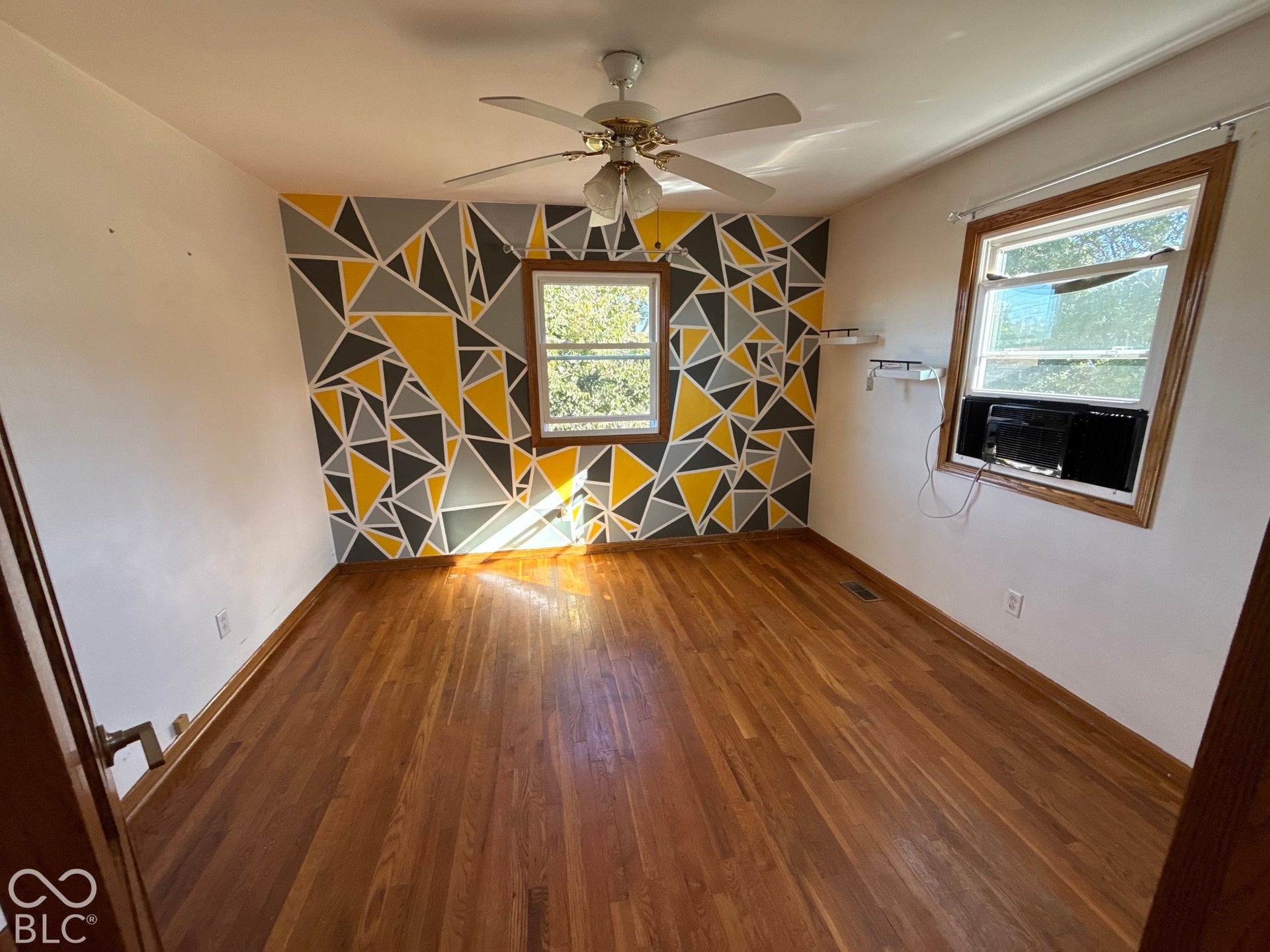 Wood-finish flooring, two windows with wood trim, ceiling fan, and a wall-mounted air conditioning unit