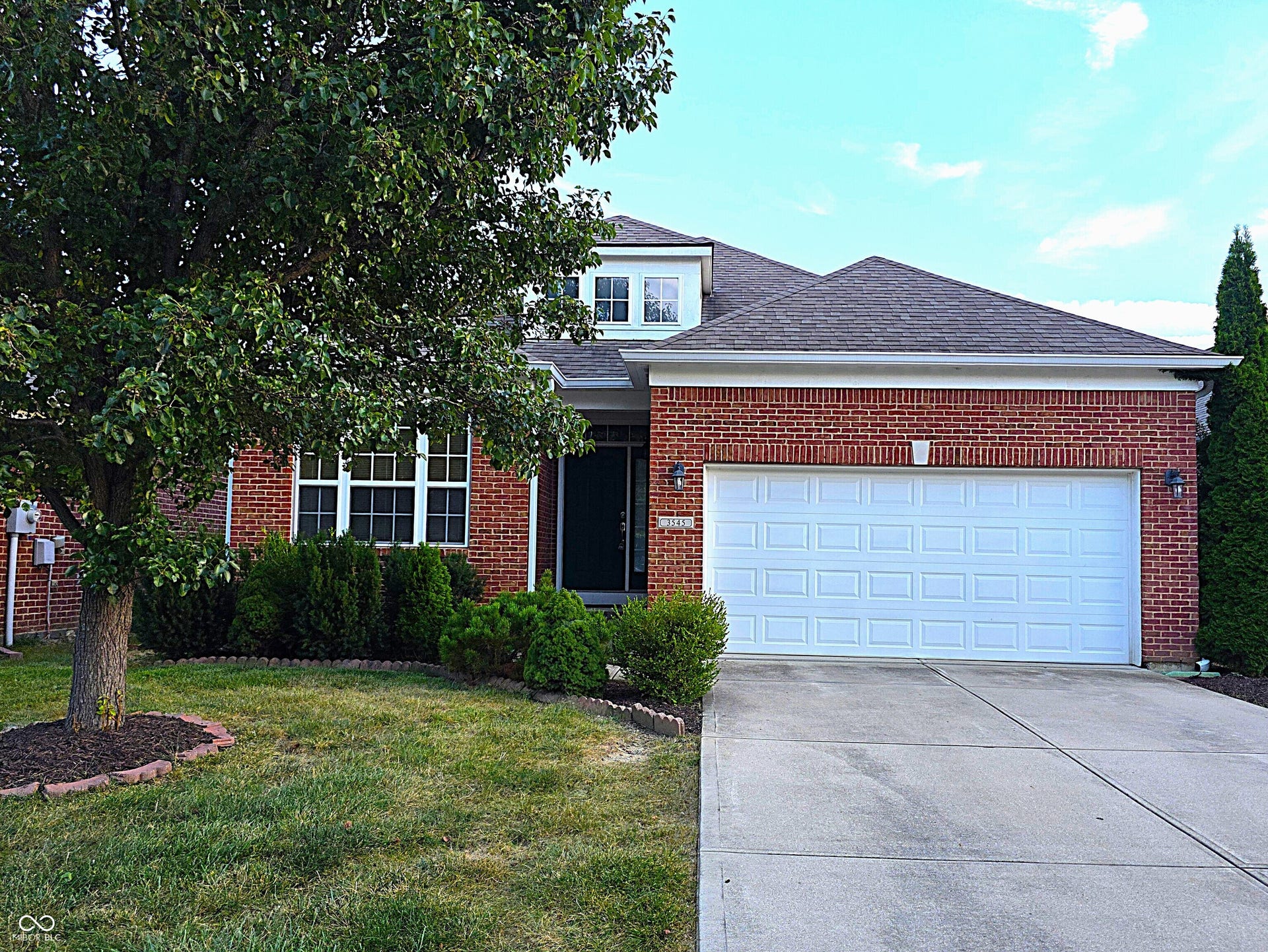 Brick facade, attached two-car garage, mature landscaping, concrete driveway, and dormer window