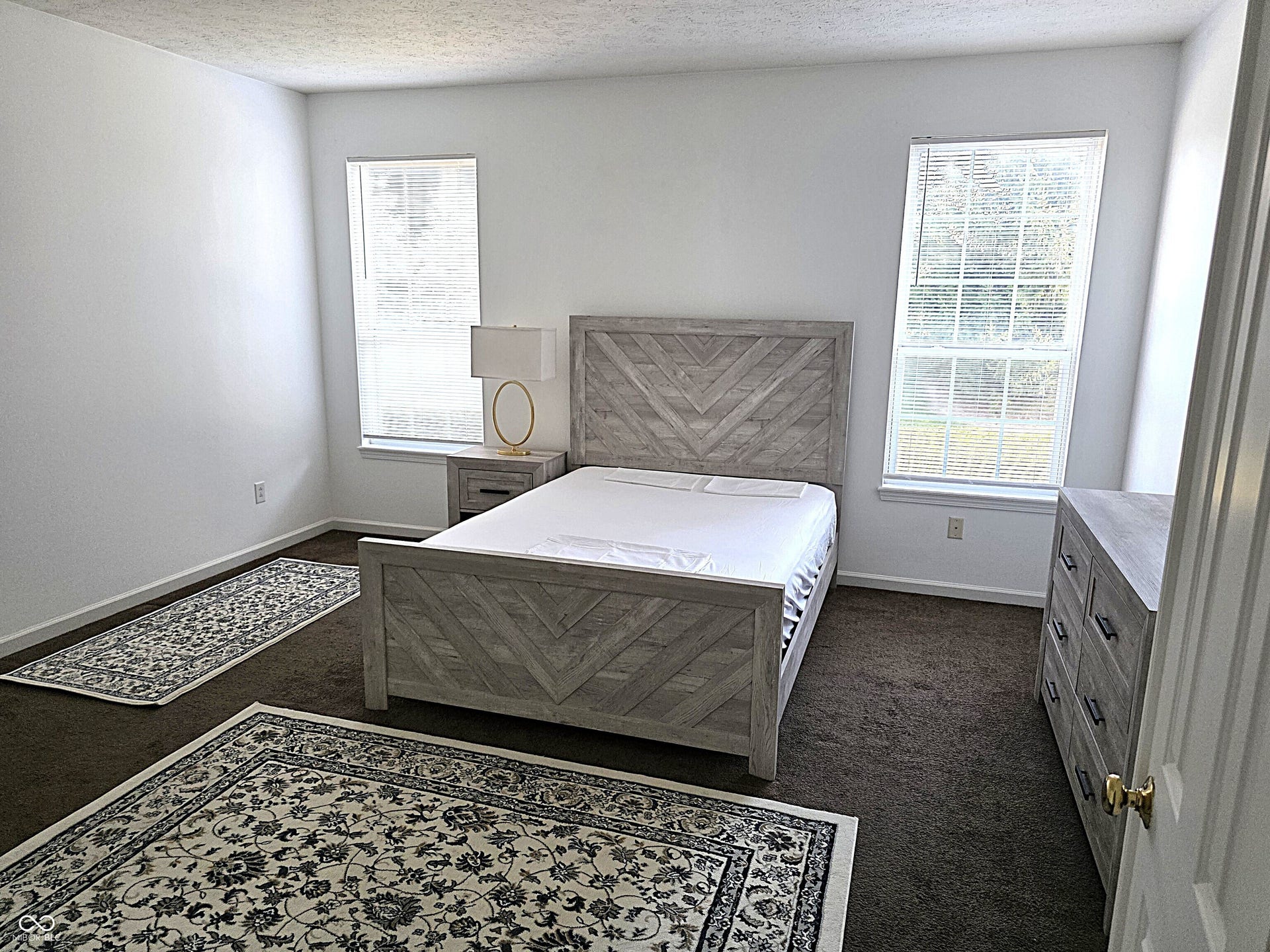 Two windows with white blinds, neutral wall paint, and dark carpet flooring