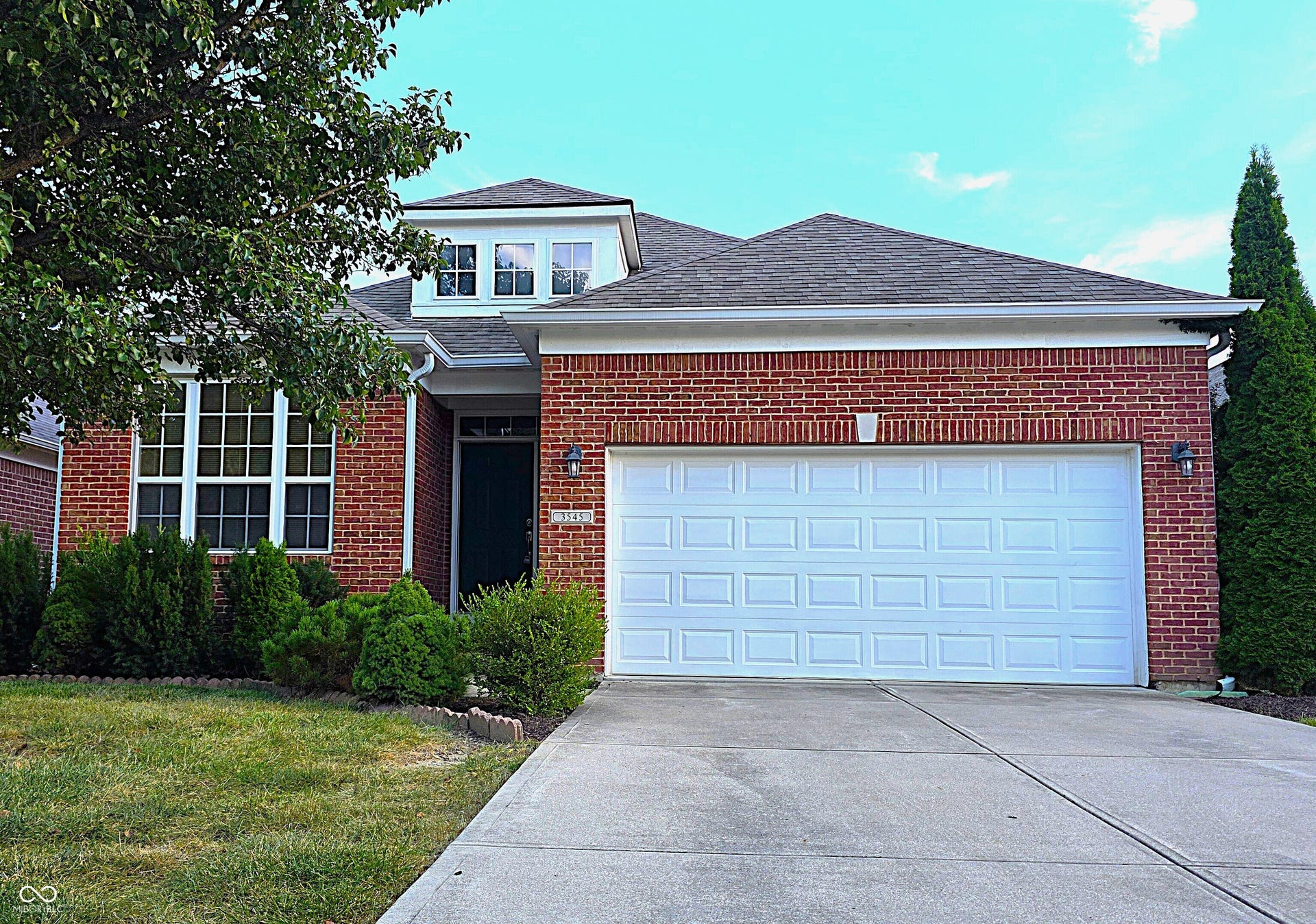 Brick exterior facade with a dormer window, an attached two-car garage, concrete driveway, and established landscaping