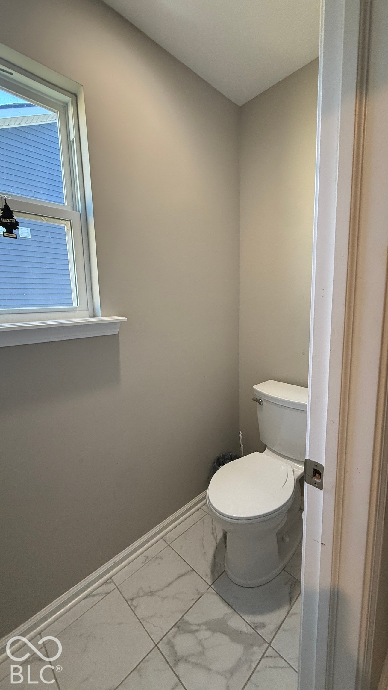 White porcelain toilet, large format floor tiling, window with white trim, and light neutral wall color