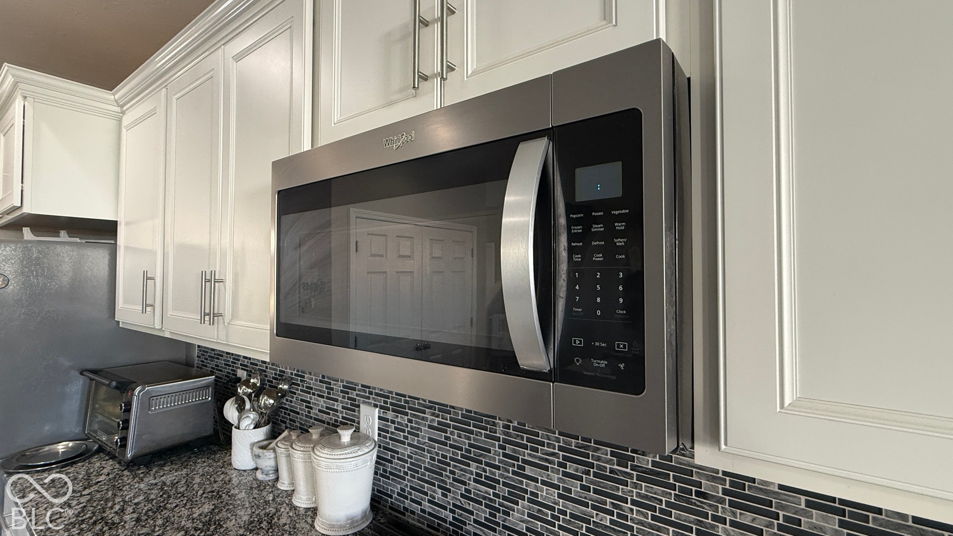 White upper cabinetry with crown molding, stainless steel microwave, black and gray mosaic tile backsplash, and dark granite countertop