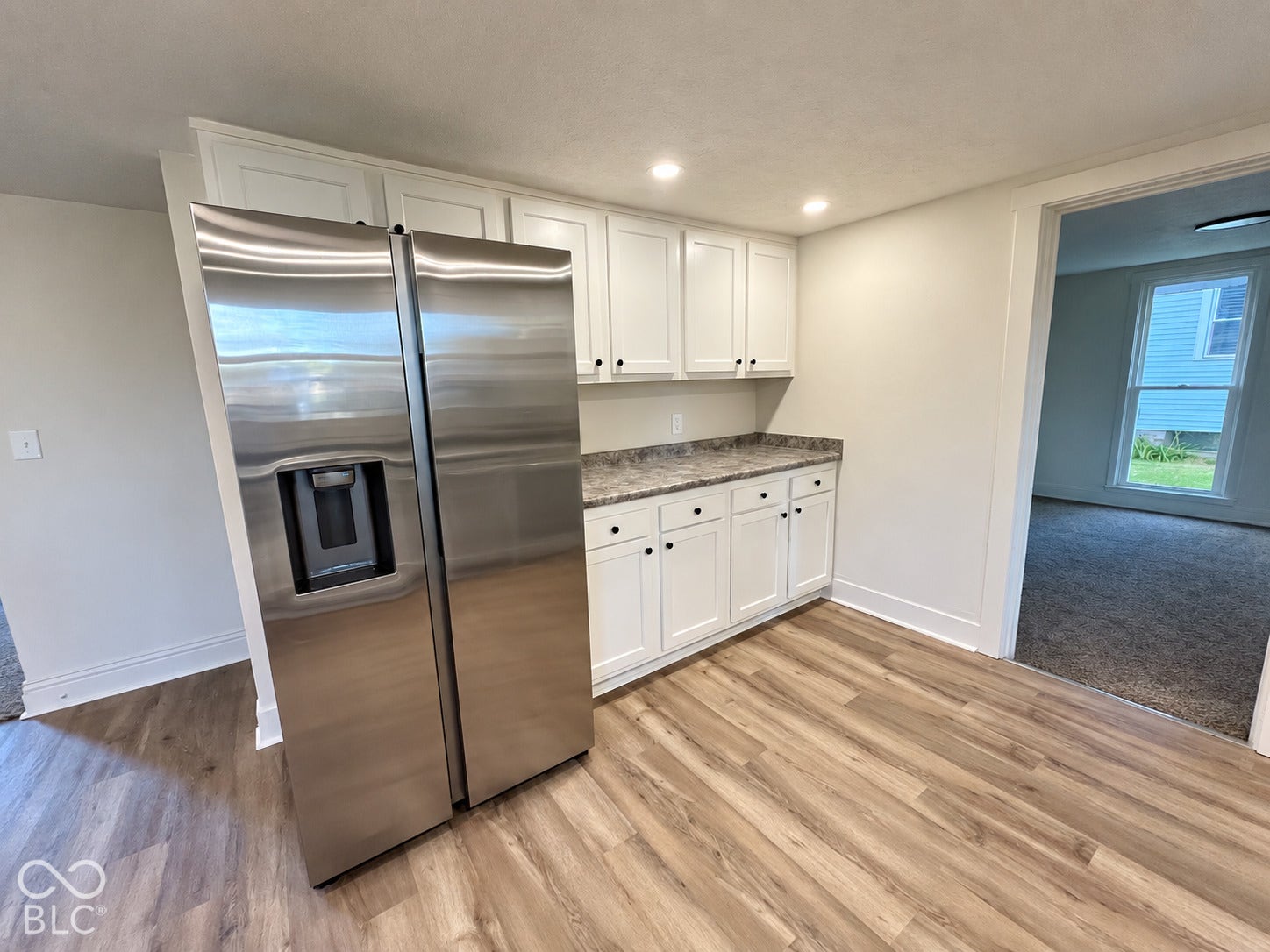 Stainless steel side-by-side refrigerator with ice and water dispenser, white shaker cabinetry with black hardware, recessed lighting, wood-finish flooring, and speckled countertops