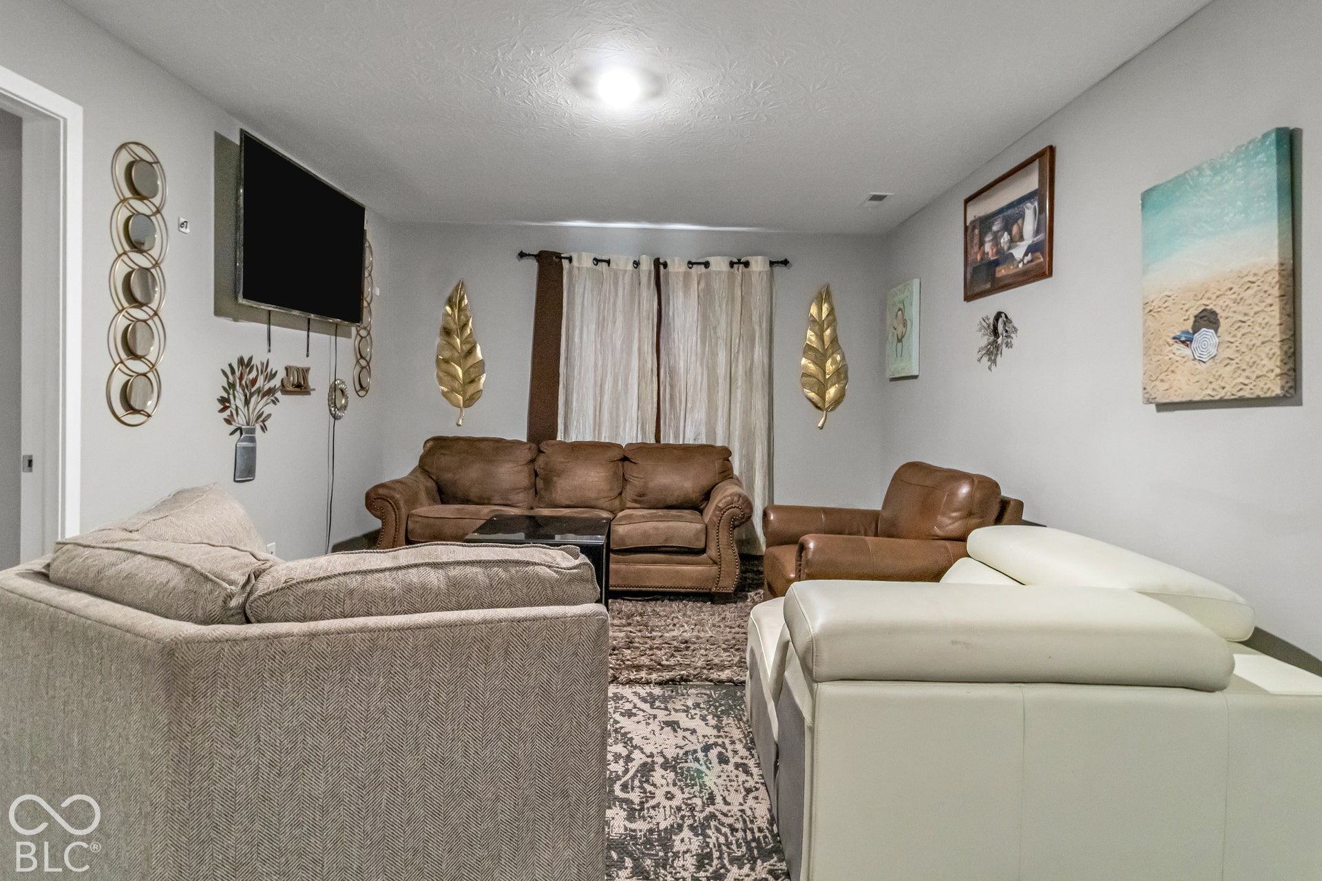 Recessed ceiling lighting illuminates a spacious room featuring light gray walls, a wall-mounted flat-screen television, and expansive window treatments