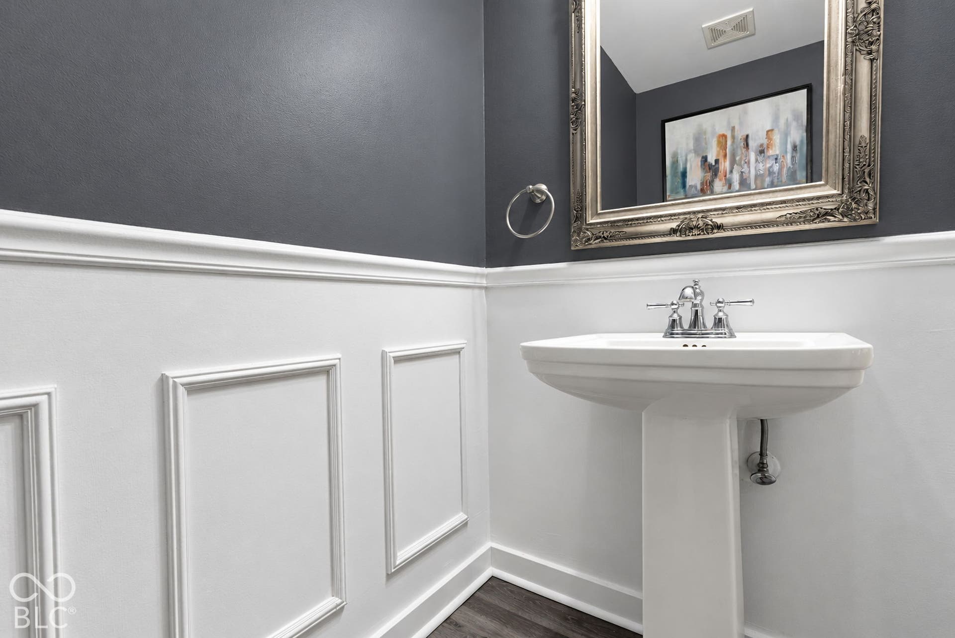 Pedestal sink with chrome fixtures, decorative wall paneling, and an ornate framed mirror
