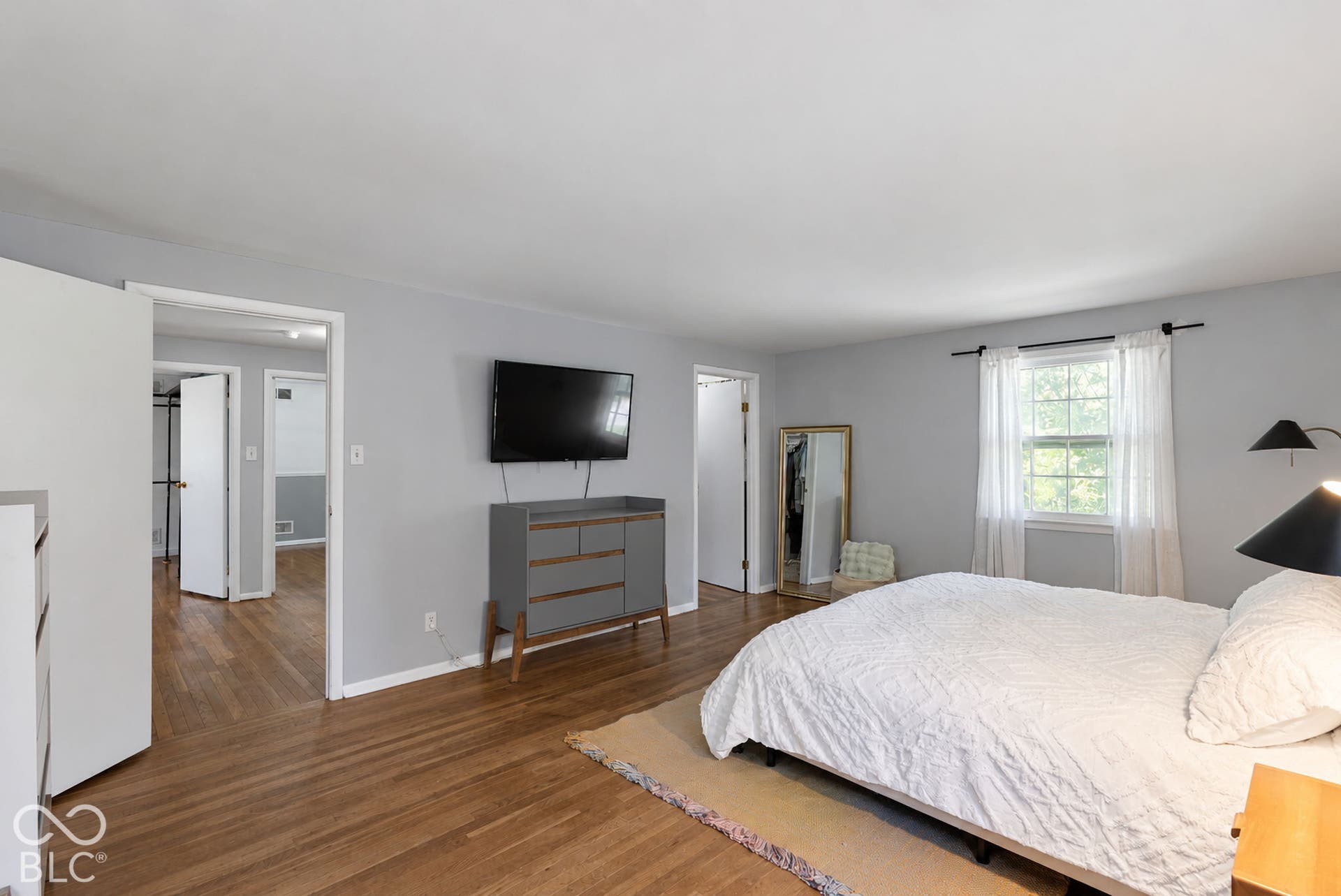 Spacious primary bedroom featuring wood-finish flooring, dual ensuite access, wall-mounted flat-screen television, built-in dresser with wood cabinetry, and abundant natural light from multiple windows