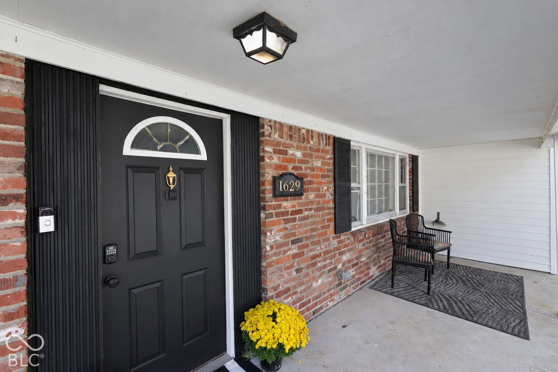 Brick exterior with a covered concrete porch, black paneled front door with a transom window, and a built-in outdoor light fixture