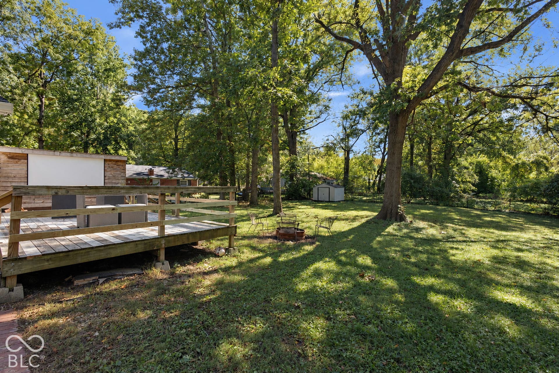 Expansive backyard featuring a wood deck with railings, mature trees, a storage shed, and a fire pit area