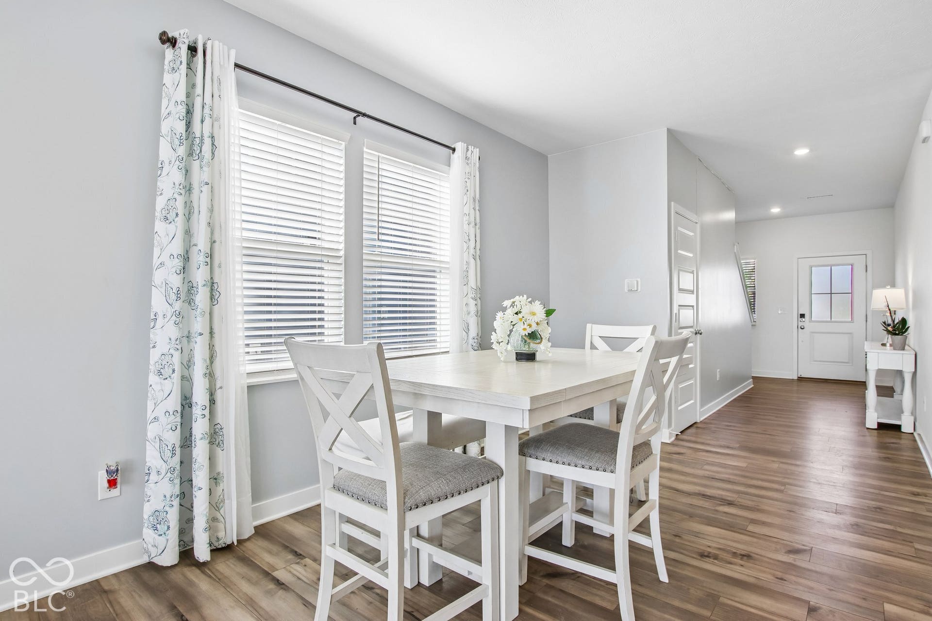 Wood-finish flooring throughout, light gray wall color, multiple windows with blinds, recessed ceiling lighting, and a paneled entry door