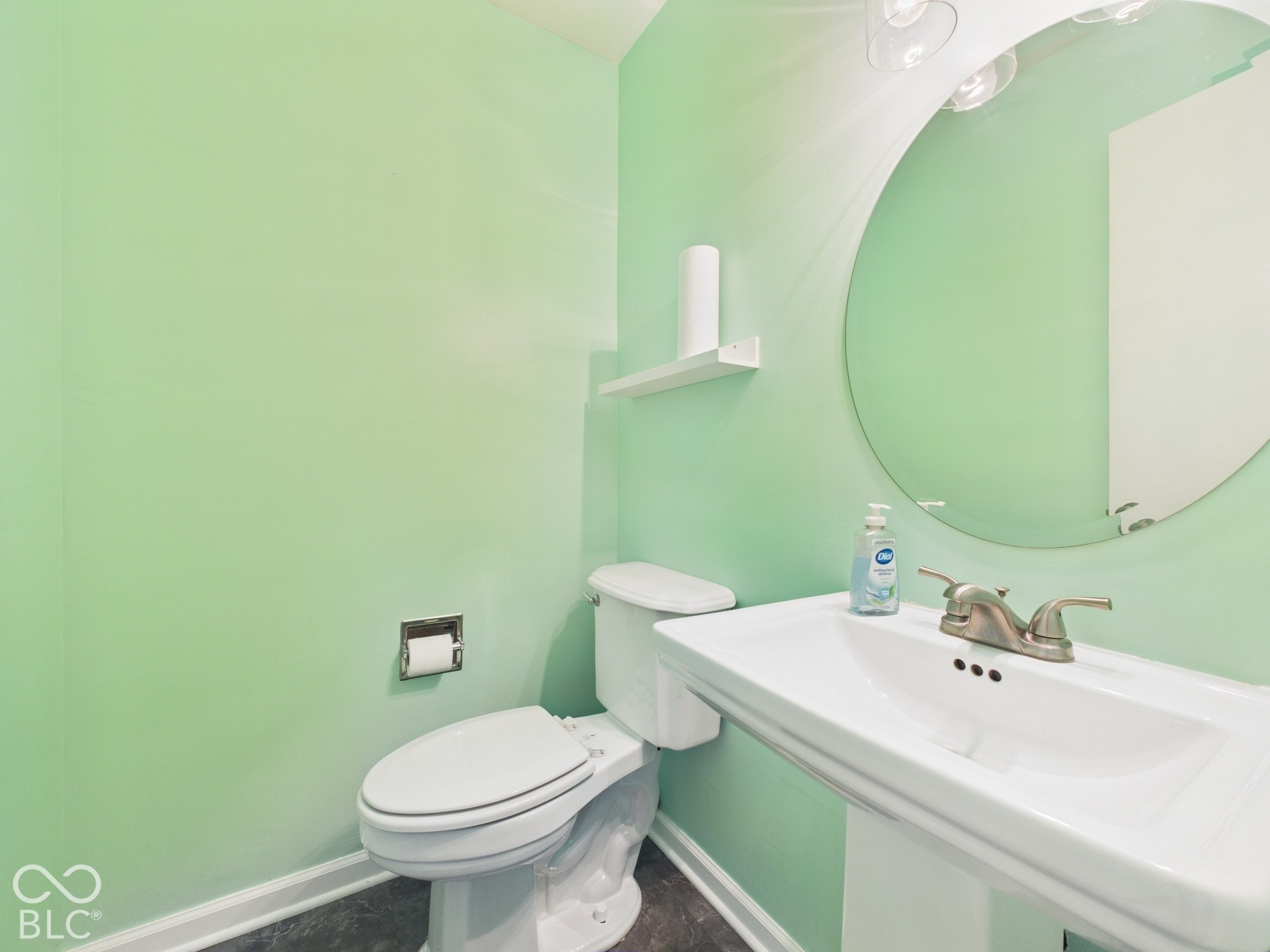Pedestal sink with brushed nickel faucet, circular wall mirror, and toilet with a chrome holder