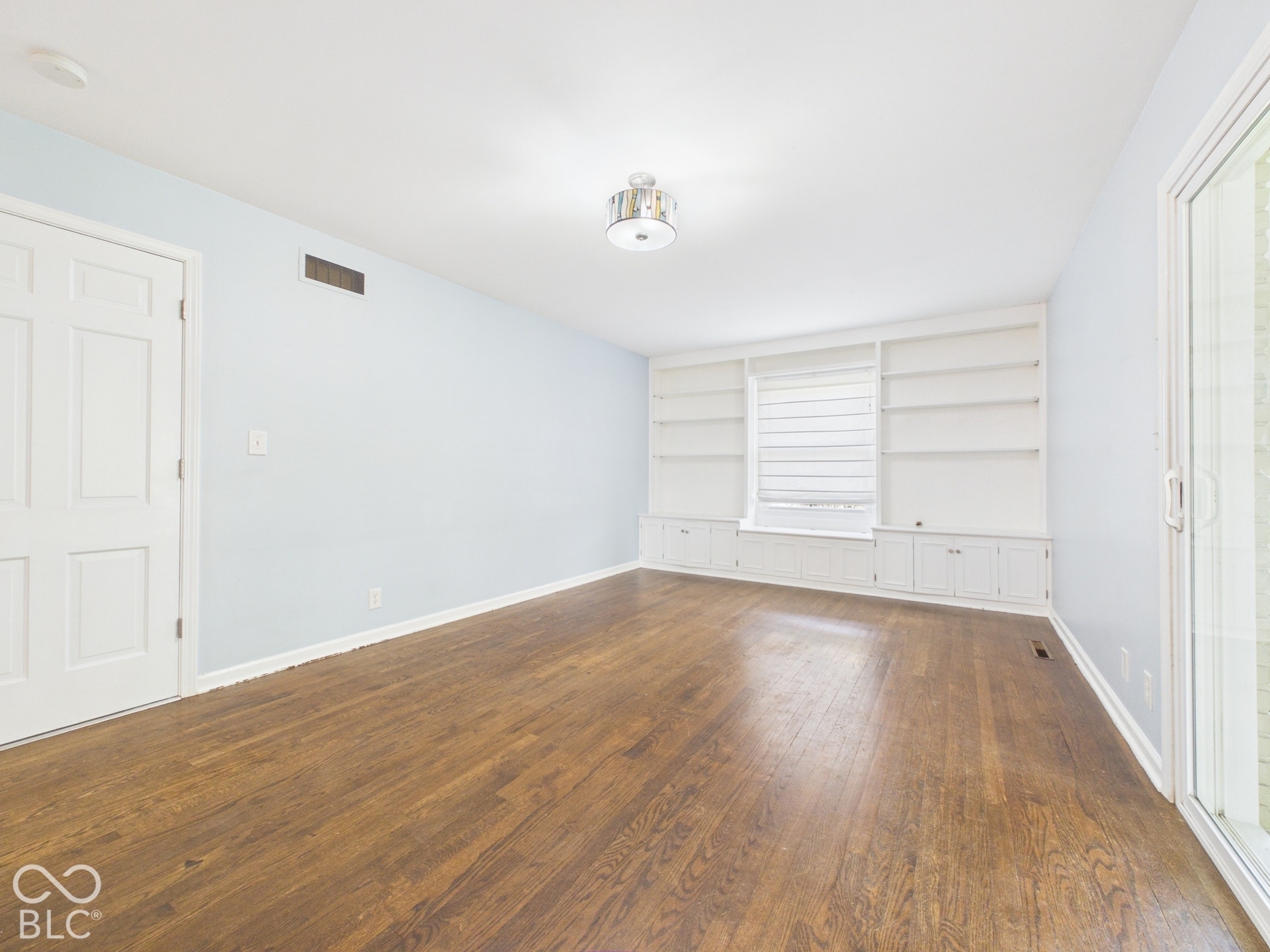 Hardwood flooring throughout, white built-in shelving with lower cabinetry, window with blind, and a sliding glass door