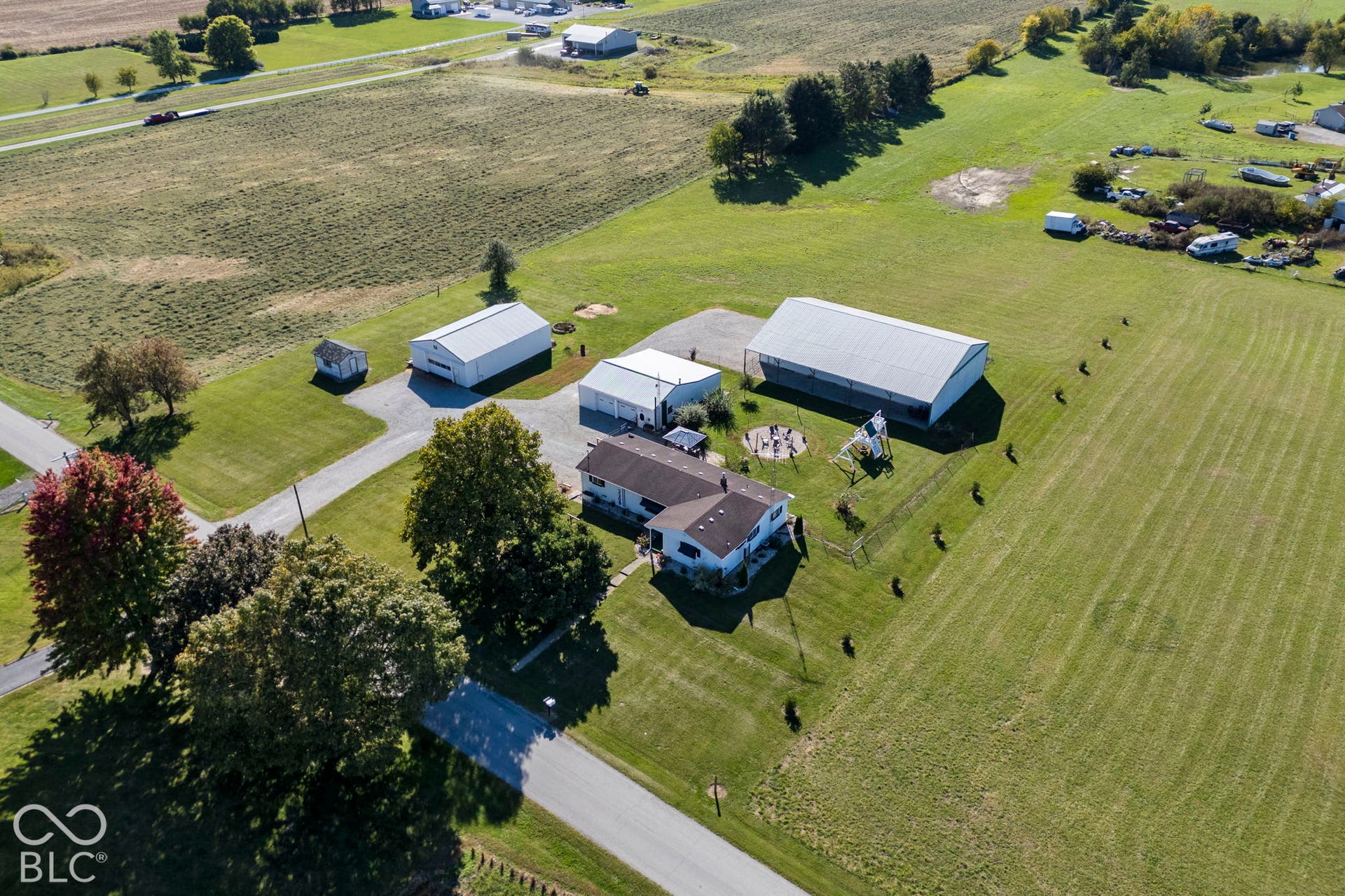 Expansive rural property featuring multiple outbuildings with white metal roofing, manicured lawn grounds with mature tree landscaping, paved driveway access, and organized equipment storage areas