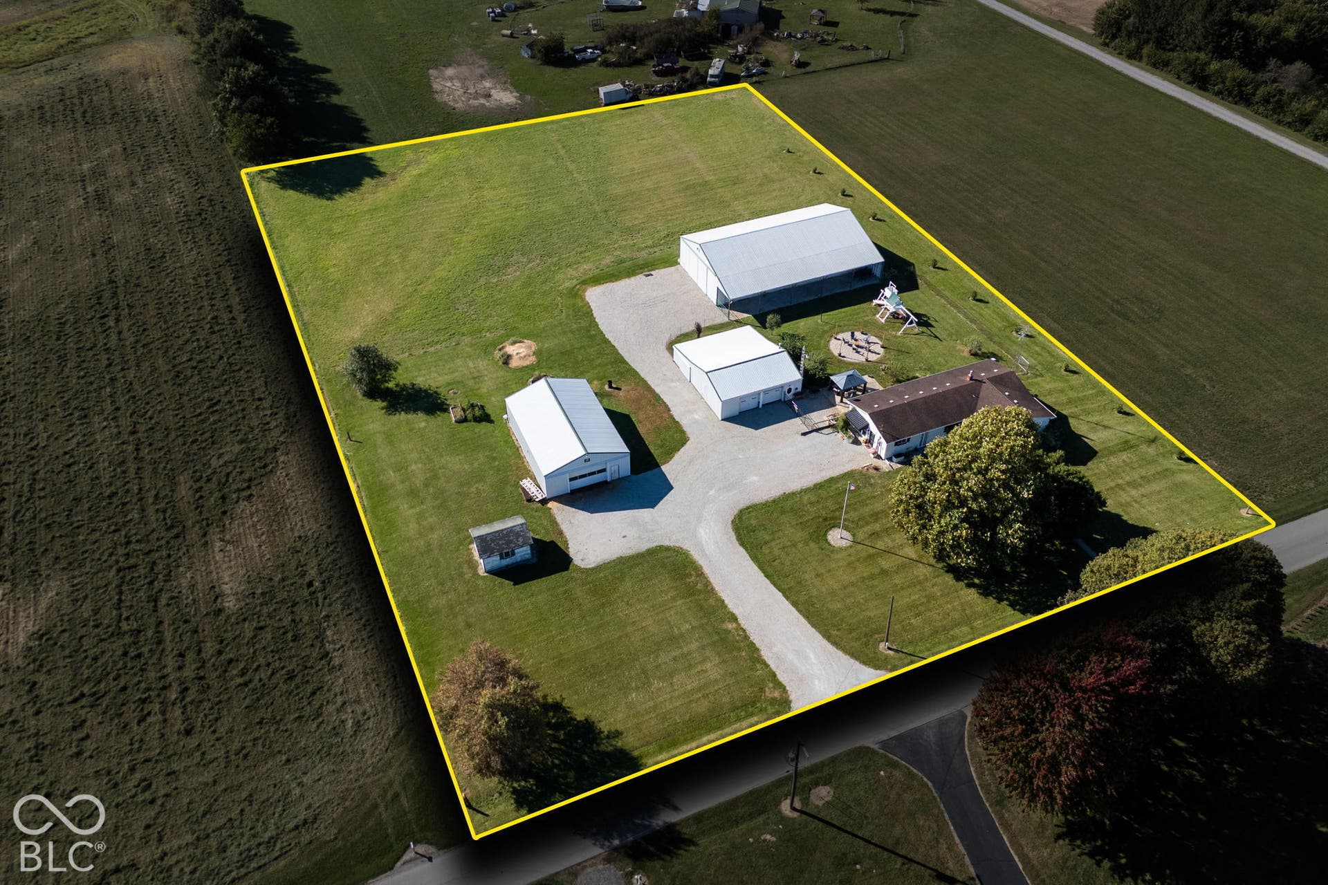 Expansive rural property featuring multiple outbuildings with metal roofing, a paved driveway with turnaround, manicured lawn grounds, mature trees providing natural screening, and substantial acreage suitable for agricultural or equestrian use