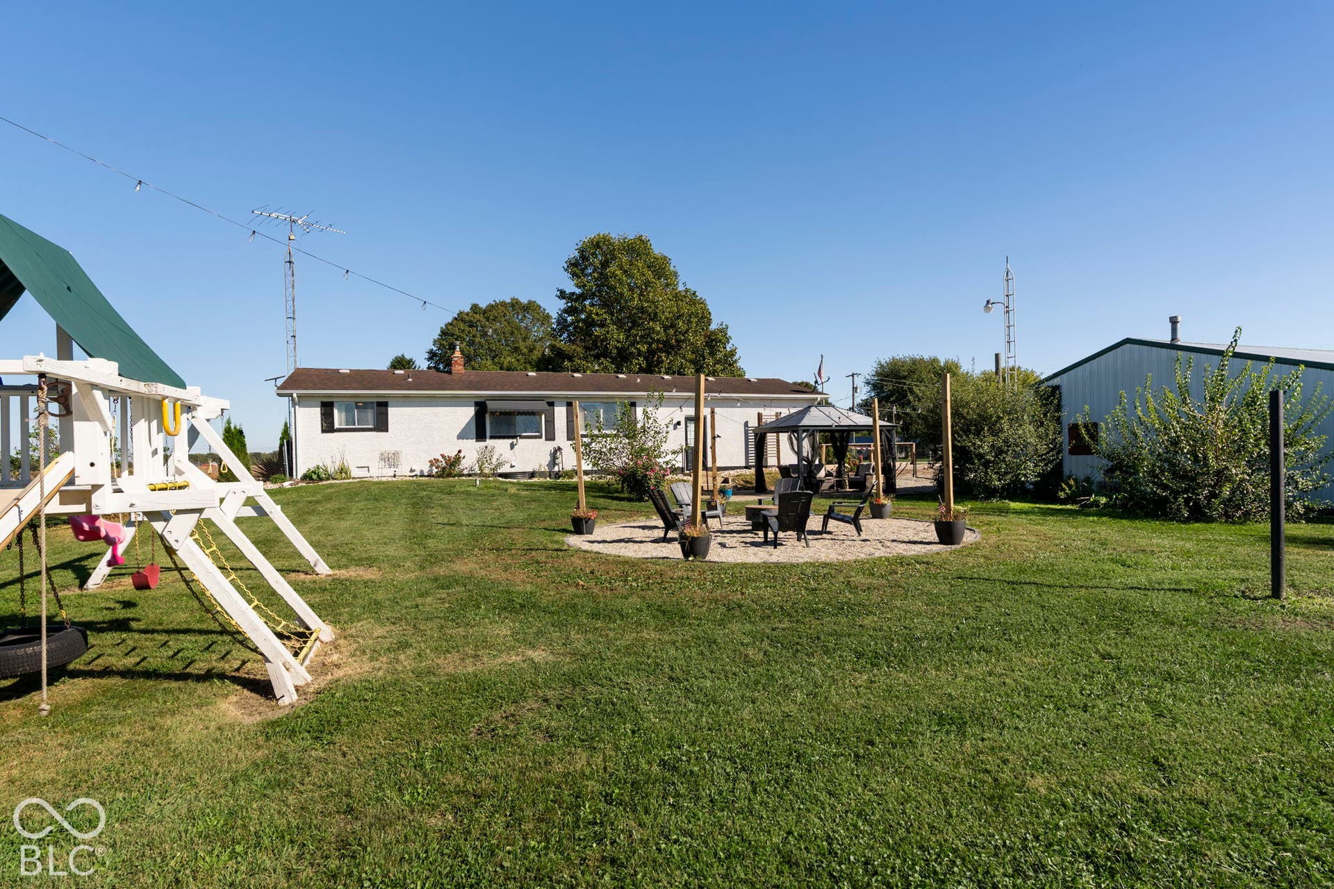 Expansive grounds featuring a manicured lawn with mature landscaping, pergola structure with seating area, single-story residential home with covered porch, and detached outbuildings