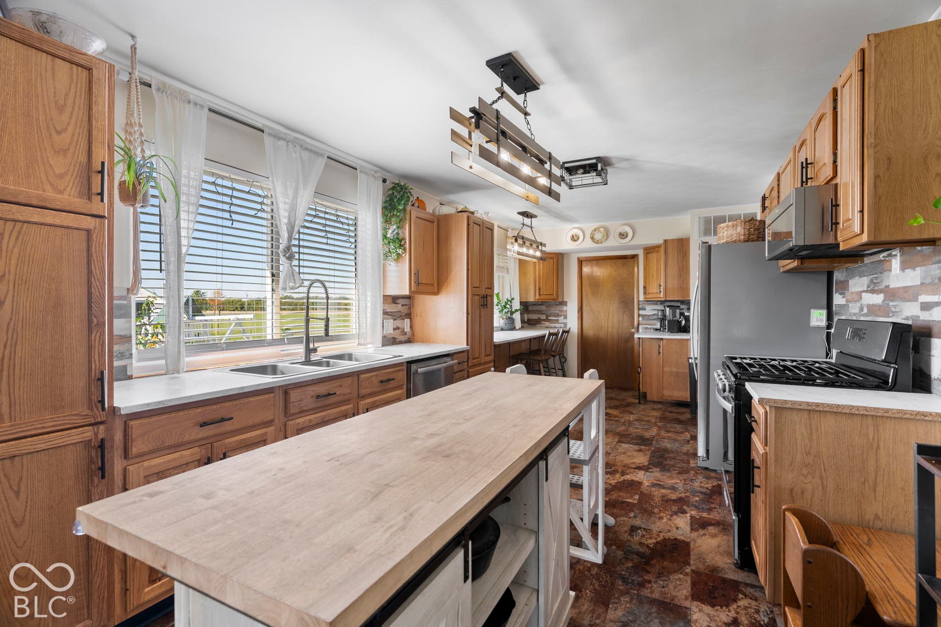 Spacious kitchen featuring wood cabinetry, a large center island with light wood-finish countertop, stainless steel gas range, undermount sink with gooseneck faucet, and abundant natural light from multi-pane windows overlooking the grounds