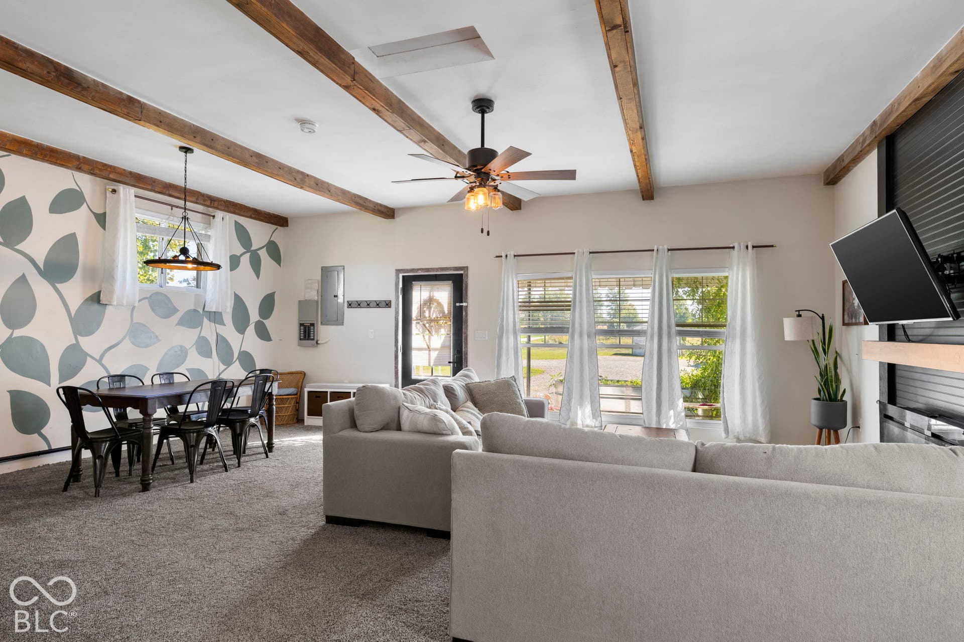 Carpeted living space featuring exposed ceiling beams, a ceiling fan with integrated lighting, multiple windows with blinds, and a built-in fireplace