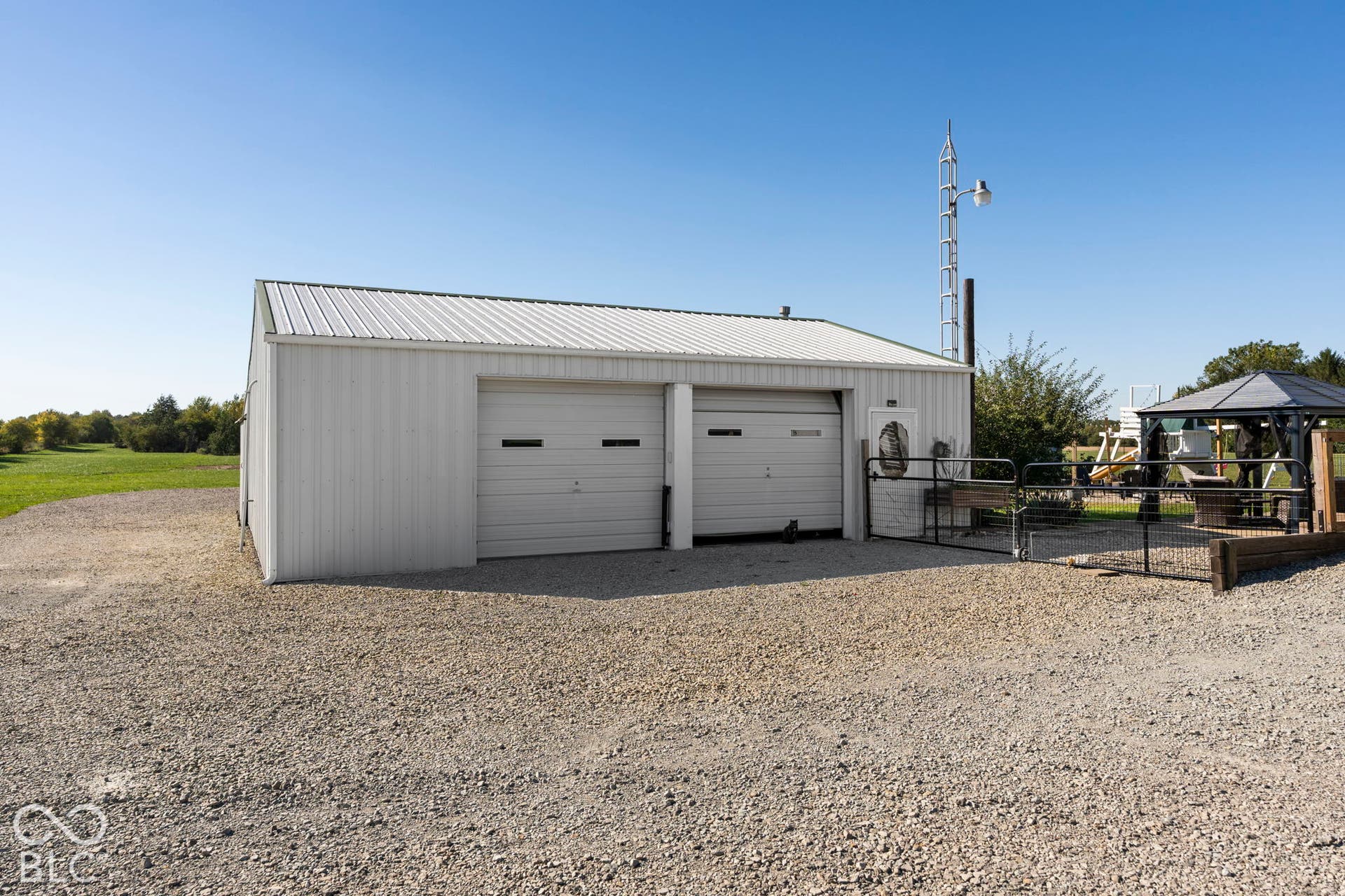 Modern metal-sided garage structure featuring two overhead bay doors, a pitched metal roof, gravel apron, and open grounds with mature landscaping and clear sky backdrop