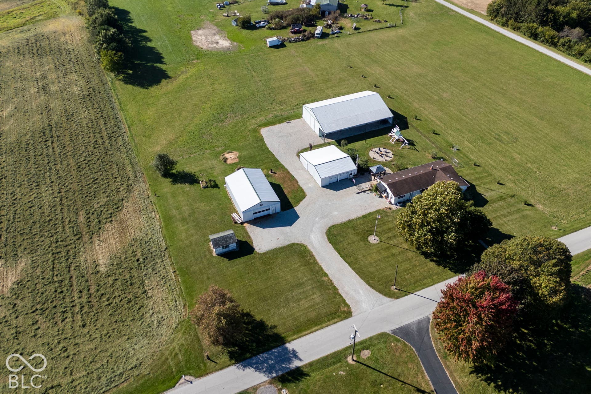 Pastoral acreage featuring multiple agricultural outbuildings with metal-frame construction, a paved driveway approach, mature landscaping with specimen trees, and expansive manicured grounds ideal for equestrian or farming operations