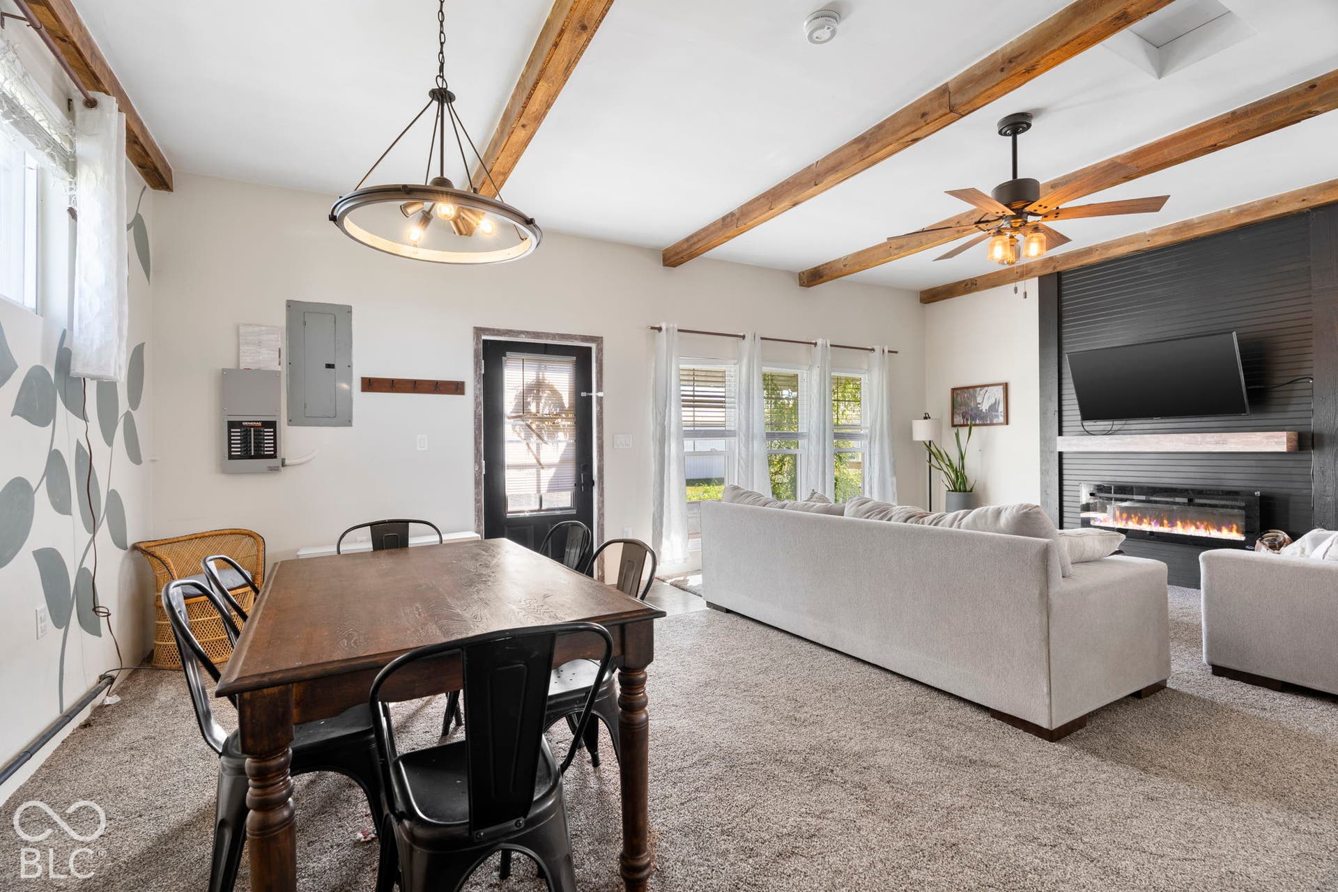 Open-concept living area featuring exposed wooden ceiling beams, a wood-finish dining table with seating for four, plush upholstered sofa, wall-mounted flat-screen television with fireplace surround, pendant lighting fixture, ceiling fan, and abundant natural light from multi-pane windows and glass doors
