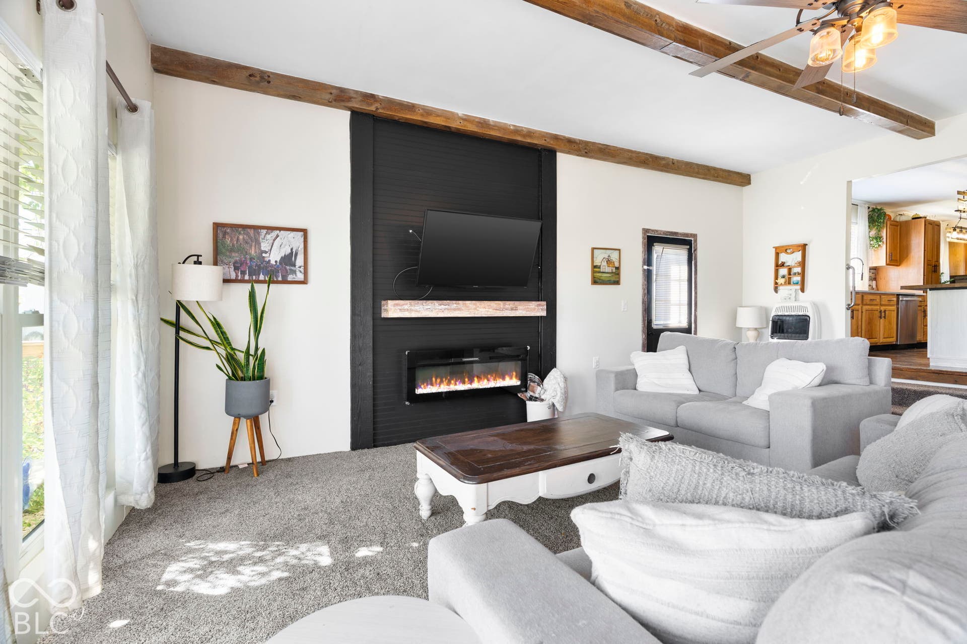 Spacious living room featuring exposed wood ceiling beams, a linear gas fireplace with black surround and hearth, expansive windows with natural light, and neutral upholstered seating areas