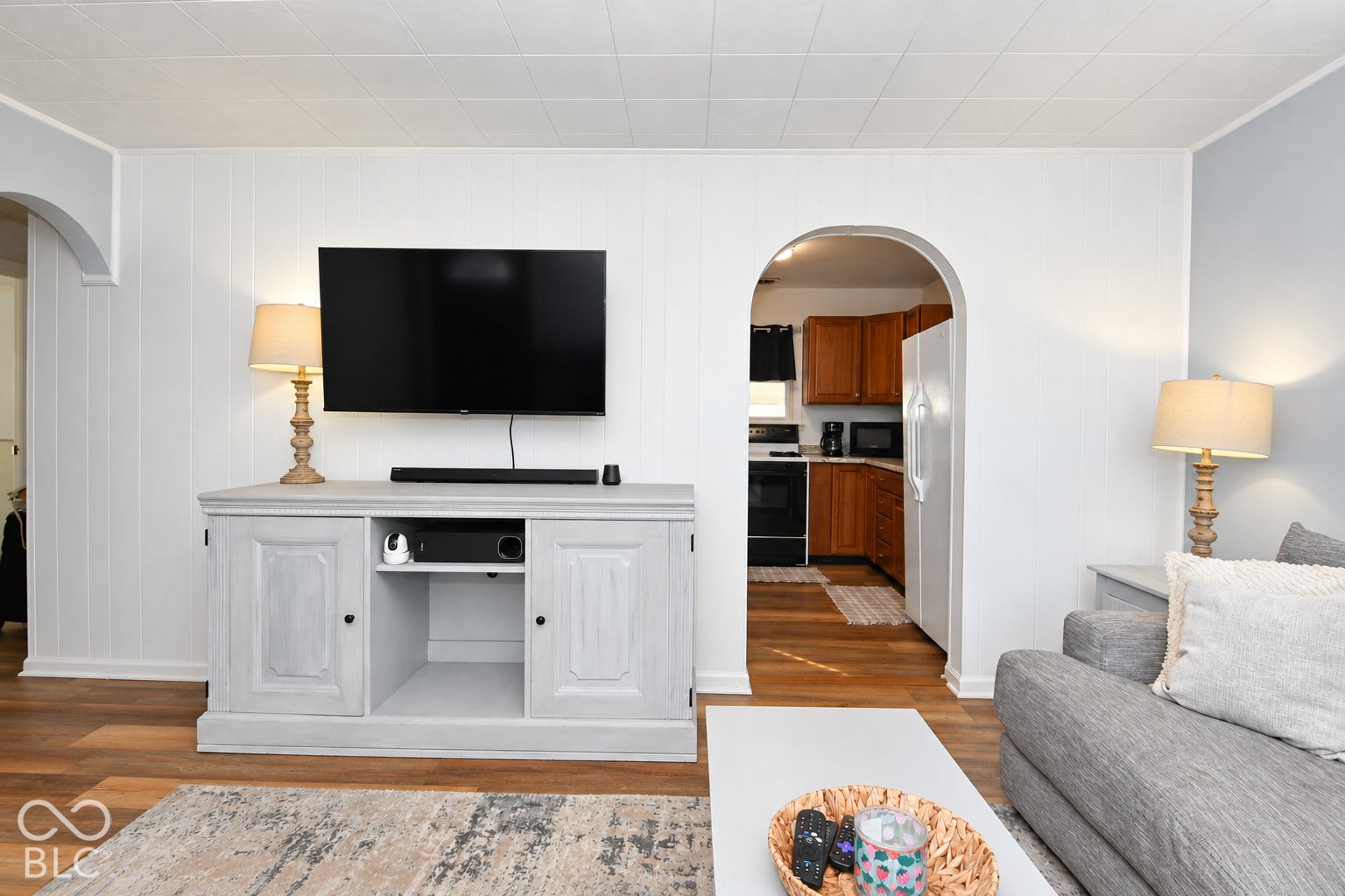 Bright primary living space featuring hardwood flooring, a white shaker-style media console with open shelving and cabinet storage, a flat-screen television with mounted soundbar, arched architectural doorway leading to additional rooms, table lamps with brass-tone bases, and neutral wall finishes throughout