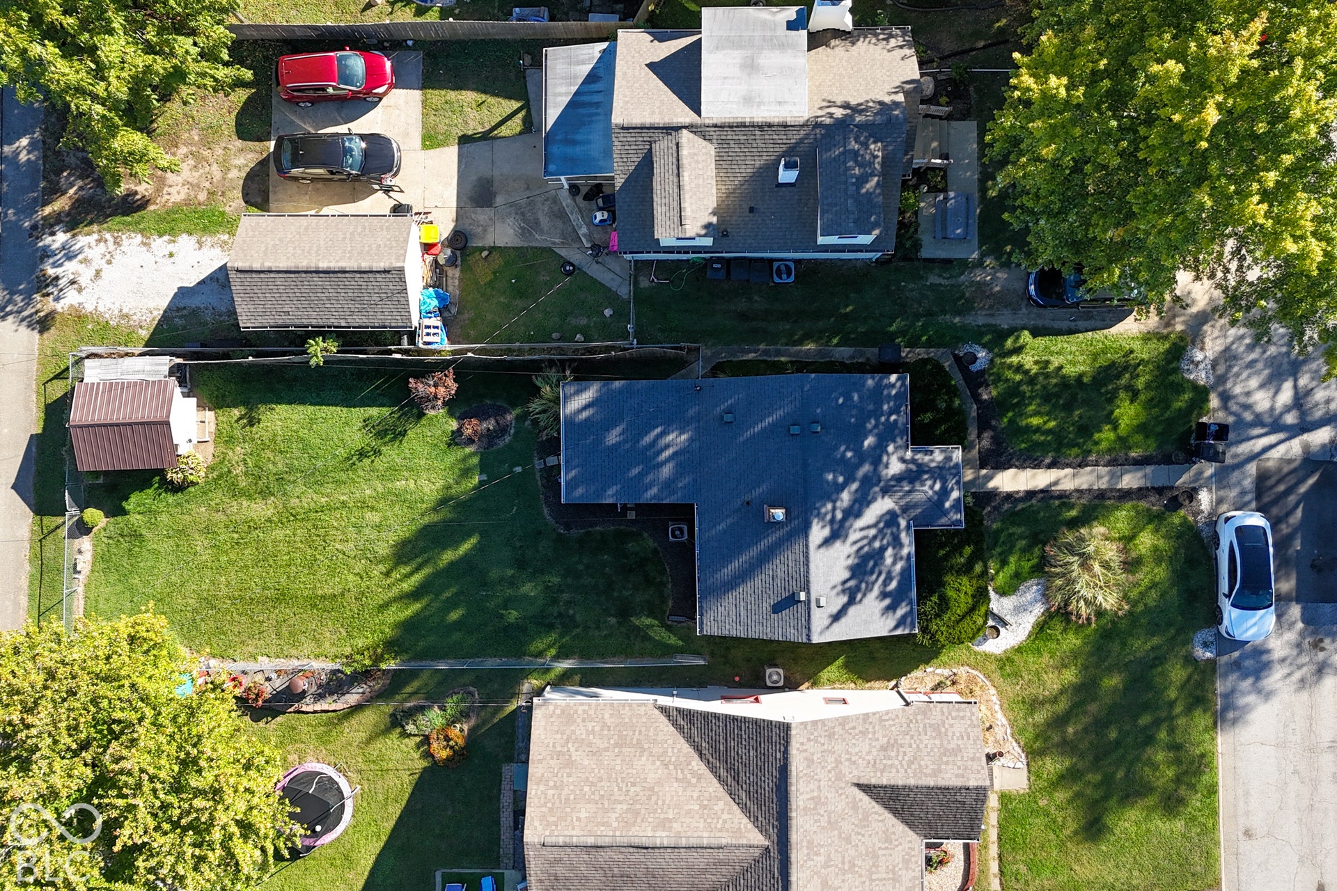 Aerial view showcasing a residential property with multiple roof sections featuring dark asphalt shingles, mature landscaping with established trees throughout the grounds, expansive lawn areas, and concrete driveway access