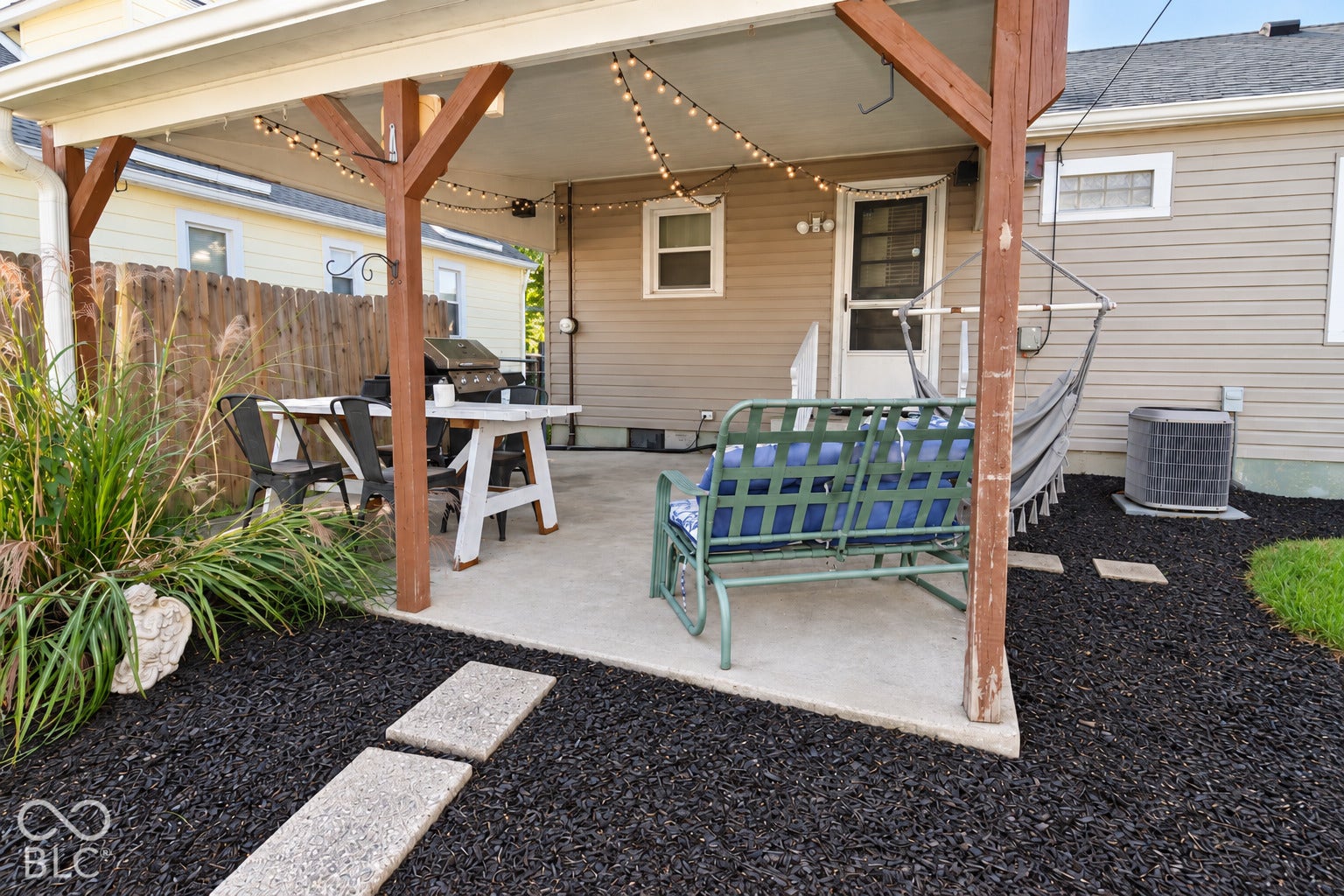 Covered patio with wooden beam construction and string lighting, complemented by black mulch landscaping, concrete stepping stones, and ornamental grasses