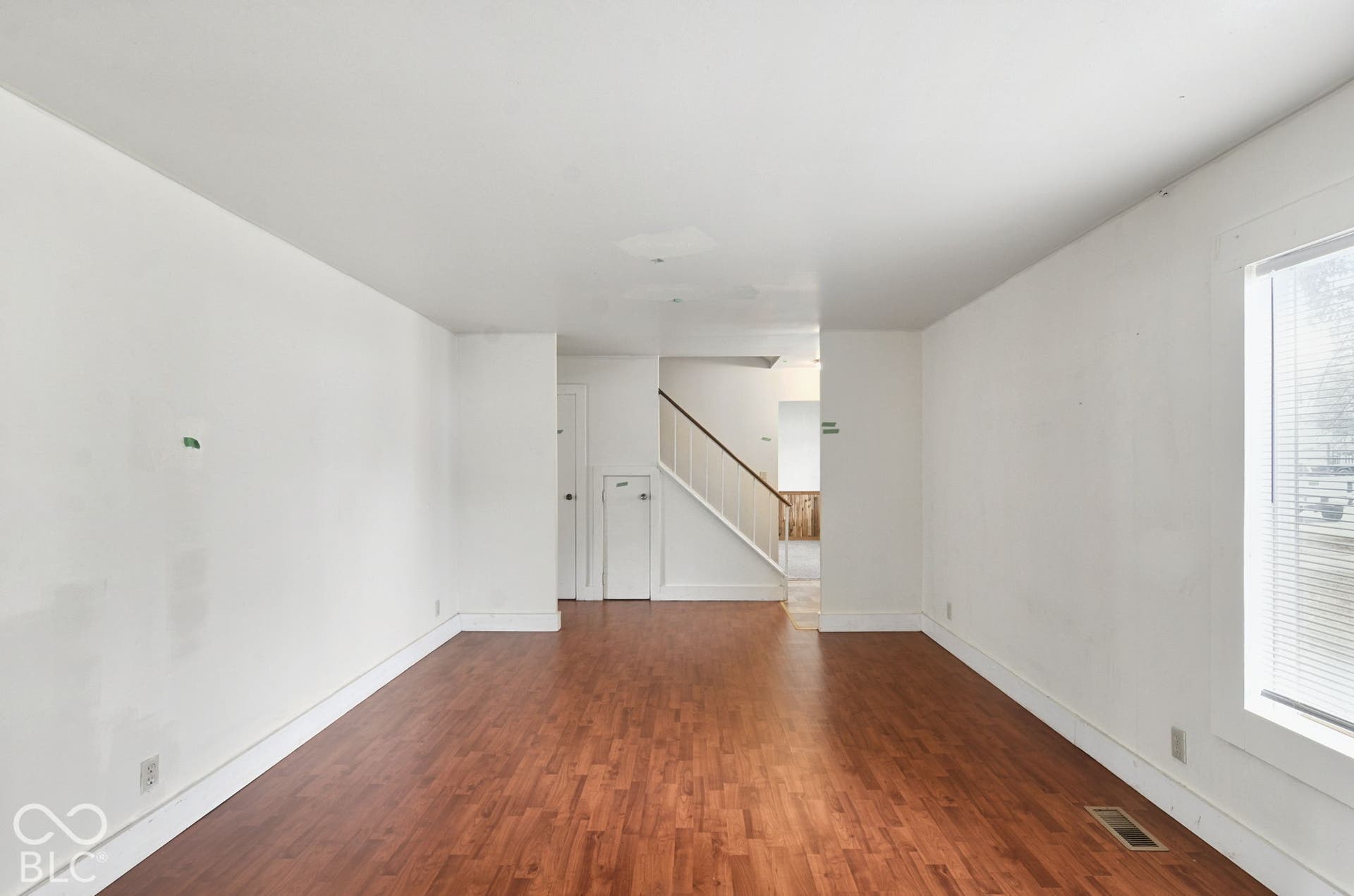 Wood-finish flooring with white baseboards, a staircase with white railings, and a window with blinds