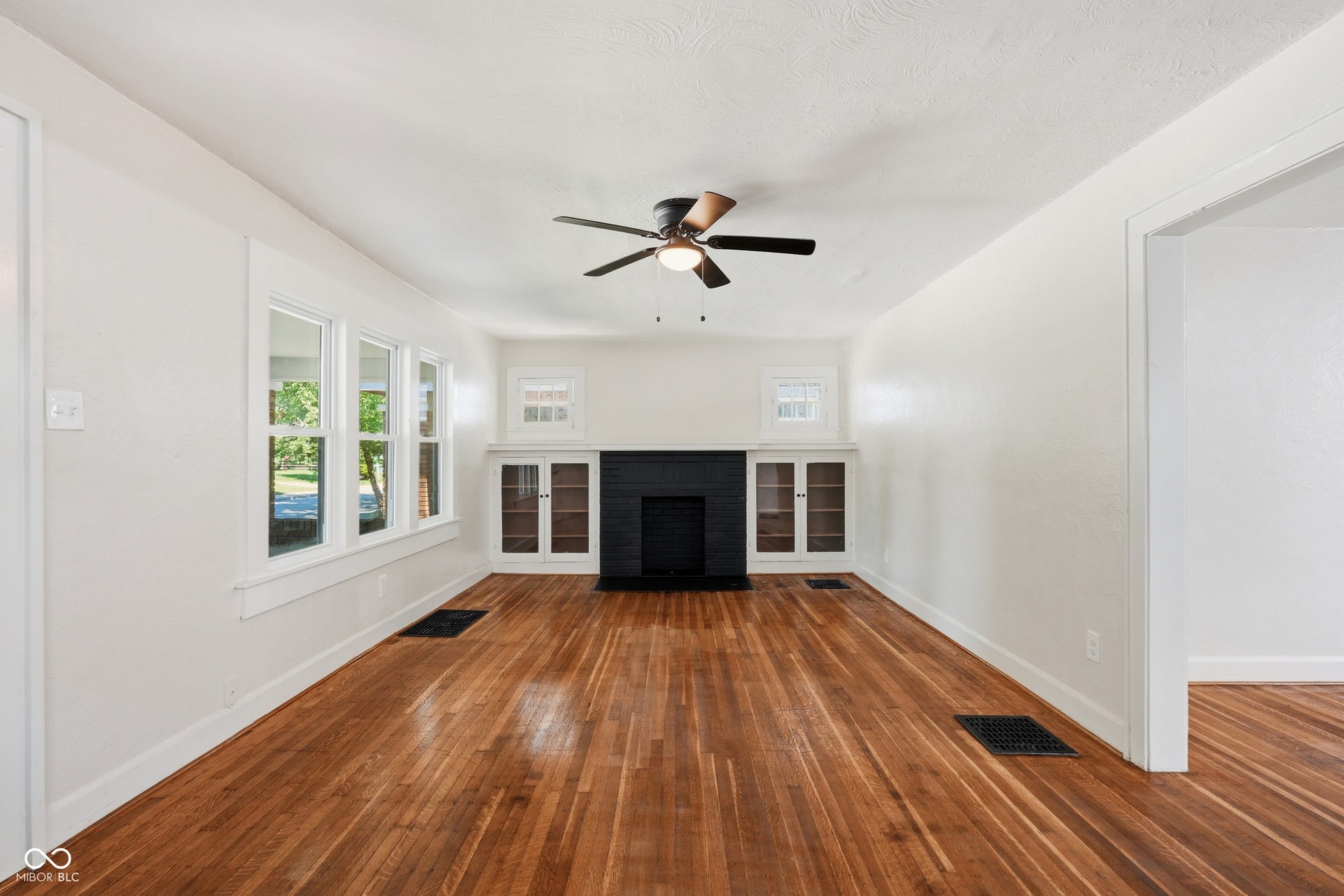 Living area featuring wood-finish flooring, a matte black fireplace surround, built-in cabinetry with glass panel doors, a ceiling fan, and multiple pane windows