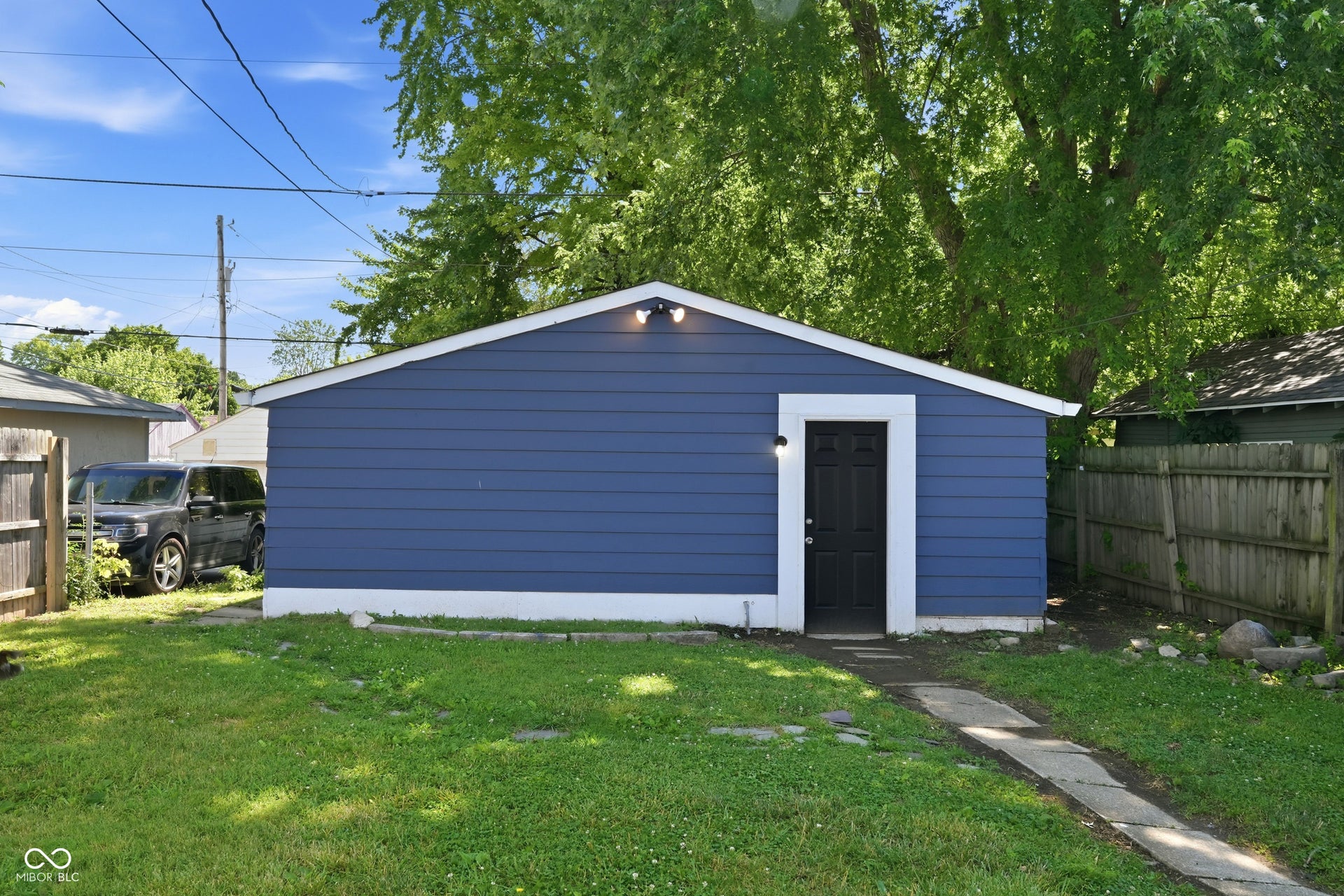 Detached garage featuring blue siding, white trim, a black entrance door with white surround, and exterior lighting