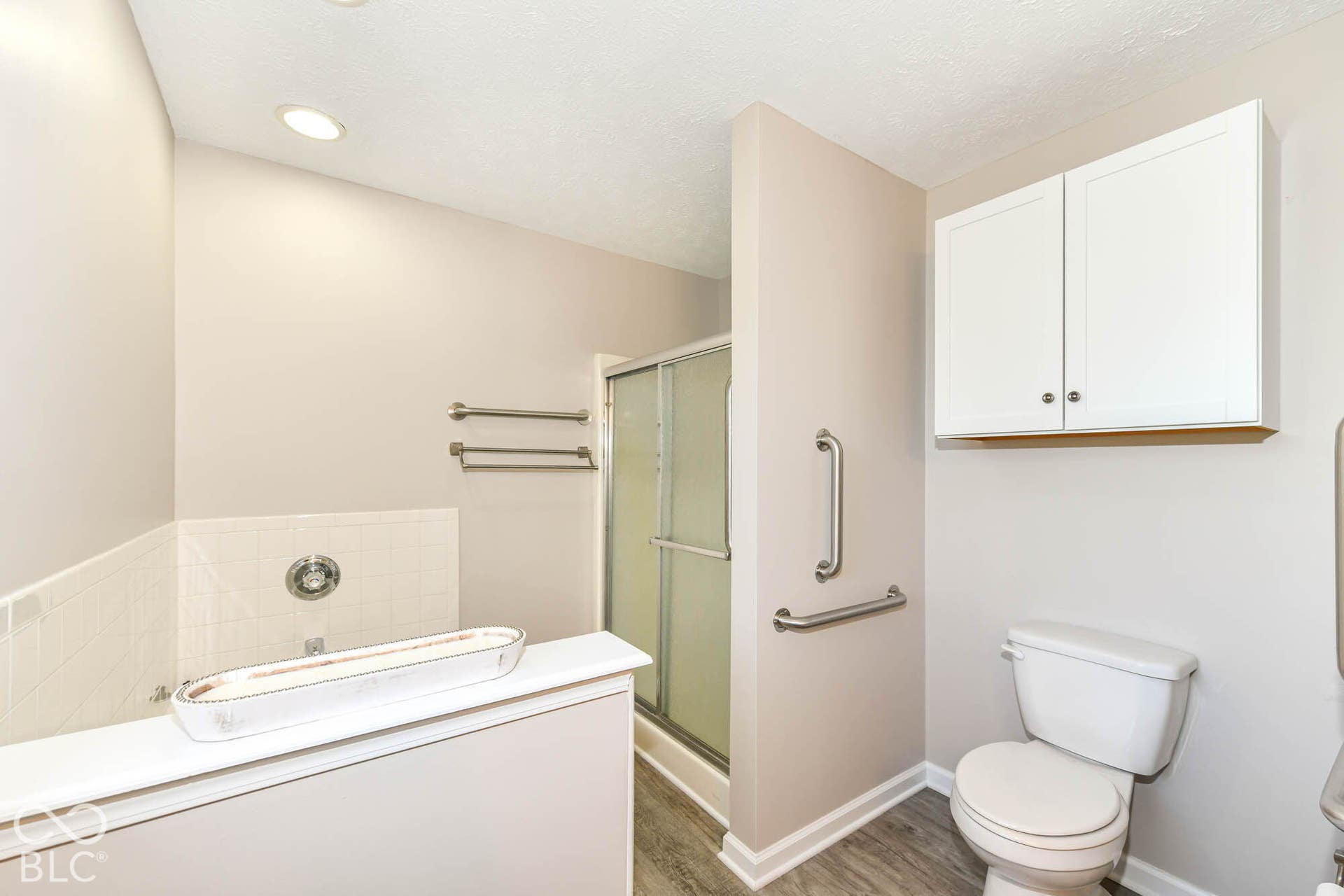 Shower with glass enclosure, white wall-mounted cabinetry, white toilet, and wood-finish flooring