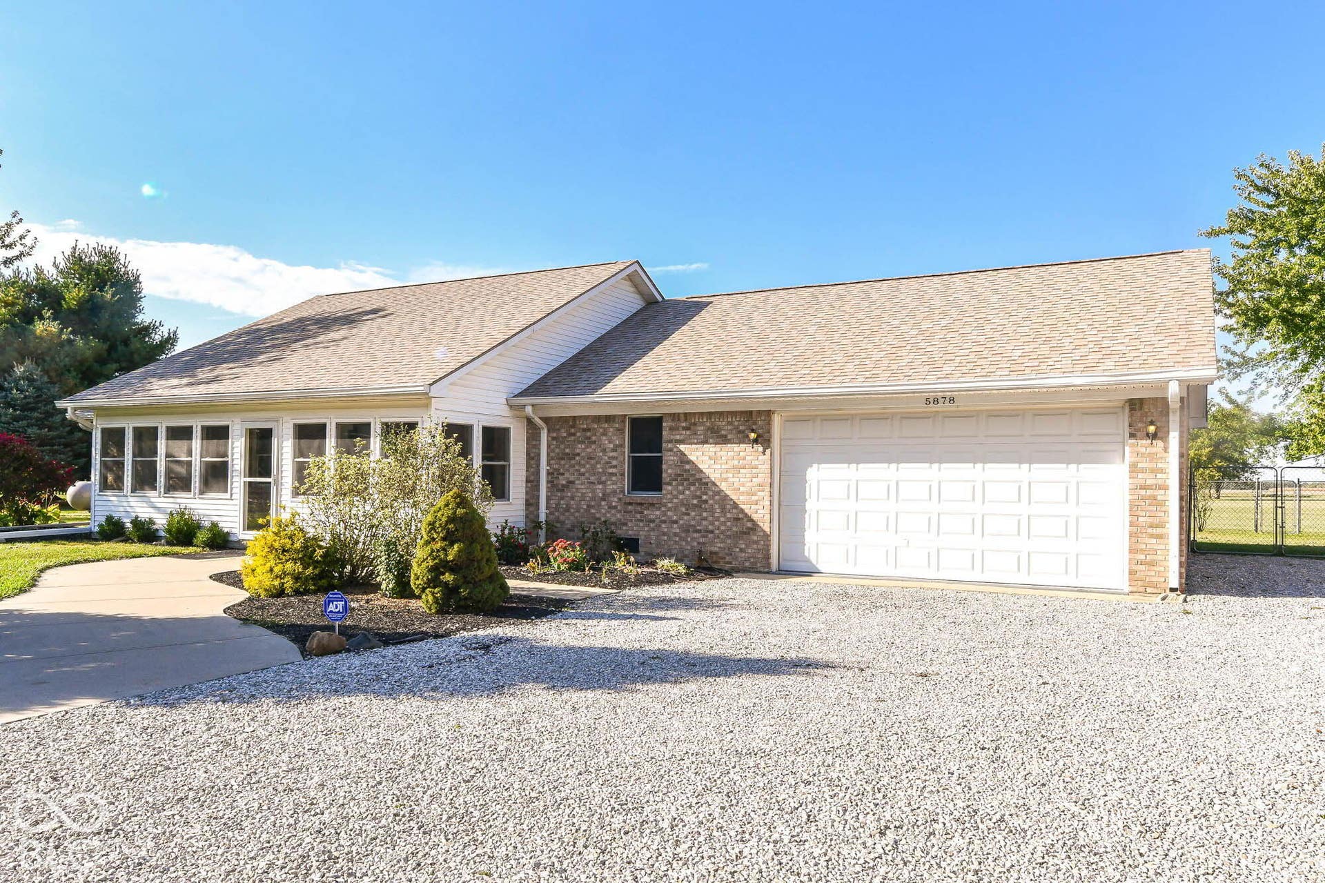 Brick and siding exterior featuring an attached two-car garage, gravel driveway, sunroom with multiple windows, and landscaped front beds
