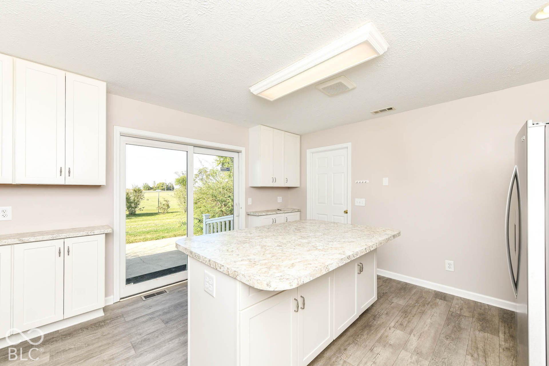 Modern kitchen featuring white cabinetry with brushed nickel hardware, a large quartz-topped island with seating, stainless steel appliances, wood-finish flooring, and abundant natural light from sliding glass doors overlooking the landscape