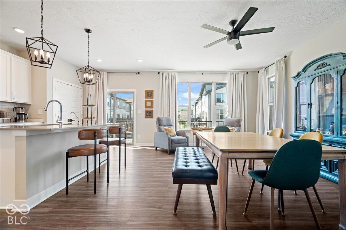 Wood-finish flooring throughout, white kitchen cabinetry with a light-colored countertop, geometric pendant lighting, ceiling fan, and a sliding glass door leading to an outdoor area
